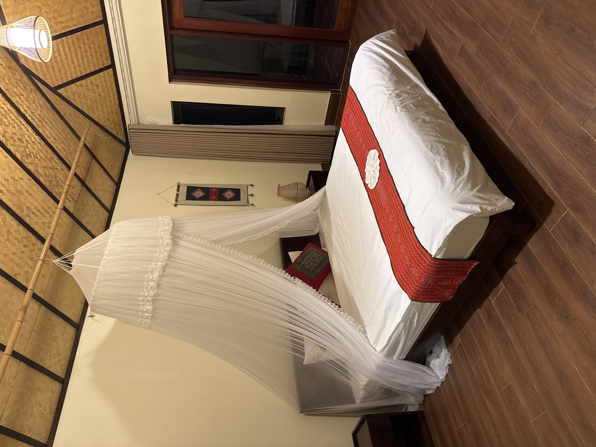 This is an interior view of a bedroom located in Ta Xua, Vietnam. The room features a high, vaulted ceiling constructed from light brown woven bamboo panels, supported by a framework of dark wooden beams. In the upper right corner, a woven pendant light fixture hangs, emitting a warm glow. The walls are painted a light cream or pale yellow color.
A large bed with a dark wooden frame dominates the center of the room. It is dressed with clean white bedding, including a white duvet and two white pillows. At the head of the bed, a decorative red pillow with a patterned central insert is positioned. A long, red patterned bed runner with white geometric designs is placed across the foot of the bed, on which a white towel is folded into a decorative shape. A white, lace-trimmed mosquito net canopy is suspended from a bamboo pole overhead, draping around the bed and partially gathered to the left, pooling slightly on the floor.
To the left of the bed, a dark wooden bedside table is visible. On the adjacent wall, a rectangular decorative textile banner with red, blue, and black geometric patterns hangs. To the right of the bed, another dark wooden bedside table holds a woven table lamp that casts a soft light. Further right, a light brown curtain partially covers a window with dark panes. Adjacent to the window are dark wooden double doors with glass panes, reflecting an indistinct outdoor or adjacent interior space. The floor is covered with dark brown, wood-look rectangular tiles arranged in a staggered pattern. No individuals are present in the image.