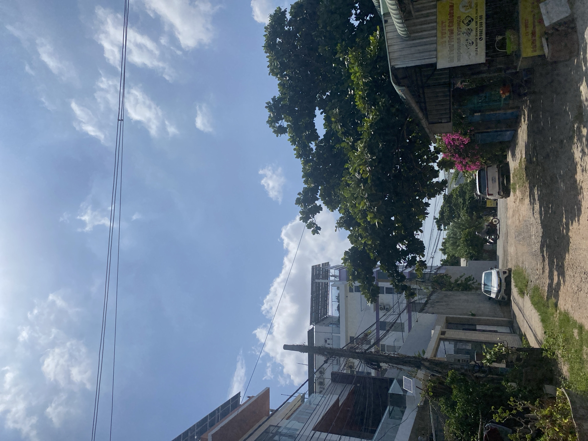 The image shows a street scene in Mũi Né, Vietnam. A white car is parked on the right side of the street, and a motorcycle is visible in the background. There are buildings on both sides of the street, with one building on the left appearing to be a multi-story structure with balconies and solar panels on the roof. A large, leafy tree overhangs the street from the right side, casting shadows on the ground. A sign with Vietnamese text and an image of chicken is attached to the side of a building on the right. The sky is blue with scattered clouds. The scene appears to be during the daytime, with bright sunlight.