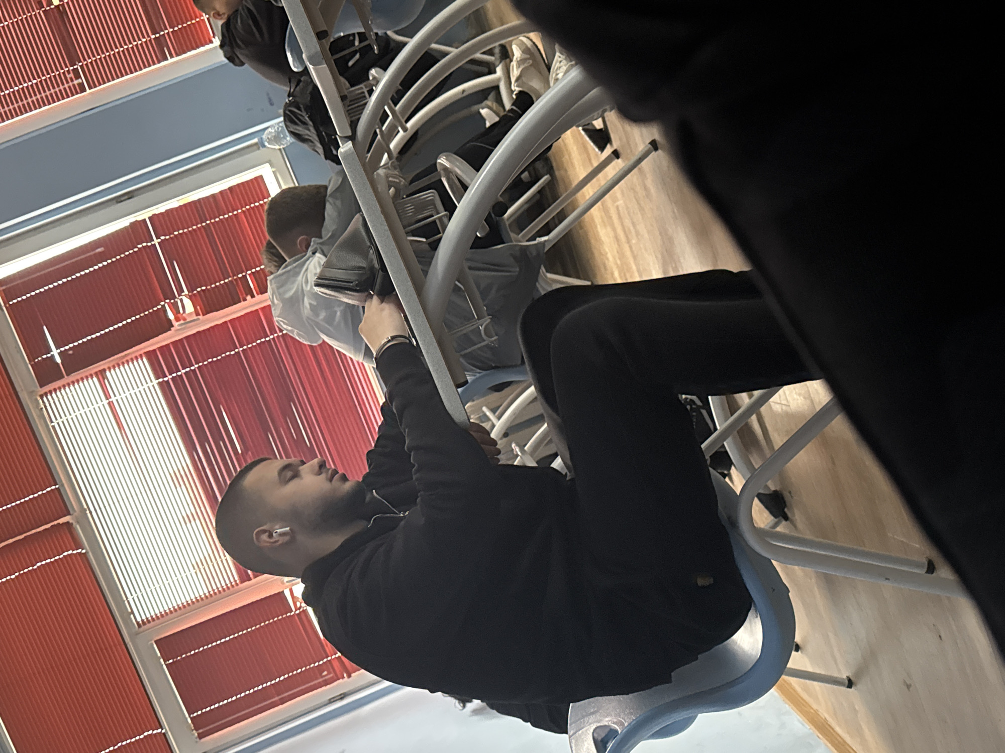 A medium shot depicts a man sitting on a blue chair in what appears to be a classroom or educational setting. He is wearing a black hoodie and black pants, with an earbud in his left ear. He is leaning forward, holding onto the metal frame of a chair in front of him. Another individual, wearing a light-colored jacket, is visible seated in the chair in front of him. Behind them, red blinds cover a window. The environment appears to be an indoor location with typical classroom furniture and decor. I cannot confirm it is in Varna, Bulgaria, based on the image.