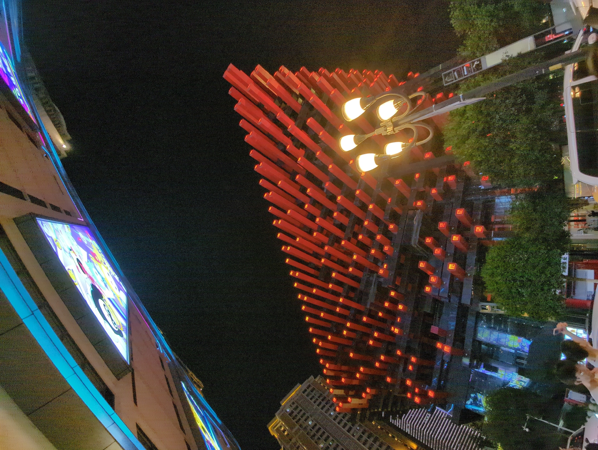 In Yuzhong District, China, at night, an architectural structure, possibly a building or a bridge element, displays vibrant blue and colorful lights against a dark sky. Below this, a multi-story building features a distinctive facade composed of numerous red, rectangular rods, each illuminated with a smaller red light at its end, creating a striking visual pattern. A streetlamp with multiple glowing bulbs stands in the foreground, its light illuminating a portion of the scene. Trees line the area, and in the lower part of the image, two individuals are visible, one taking a photograph with a phone. A white van and other vehicles are also present, suggesting an urban street setting.