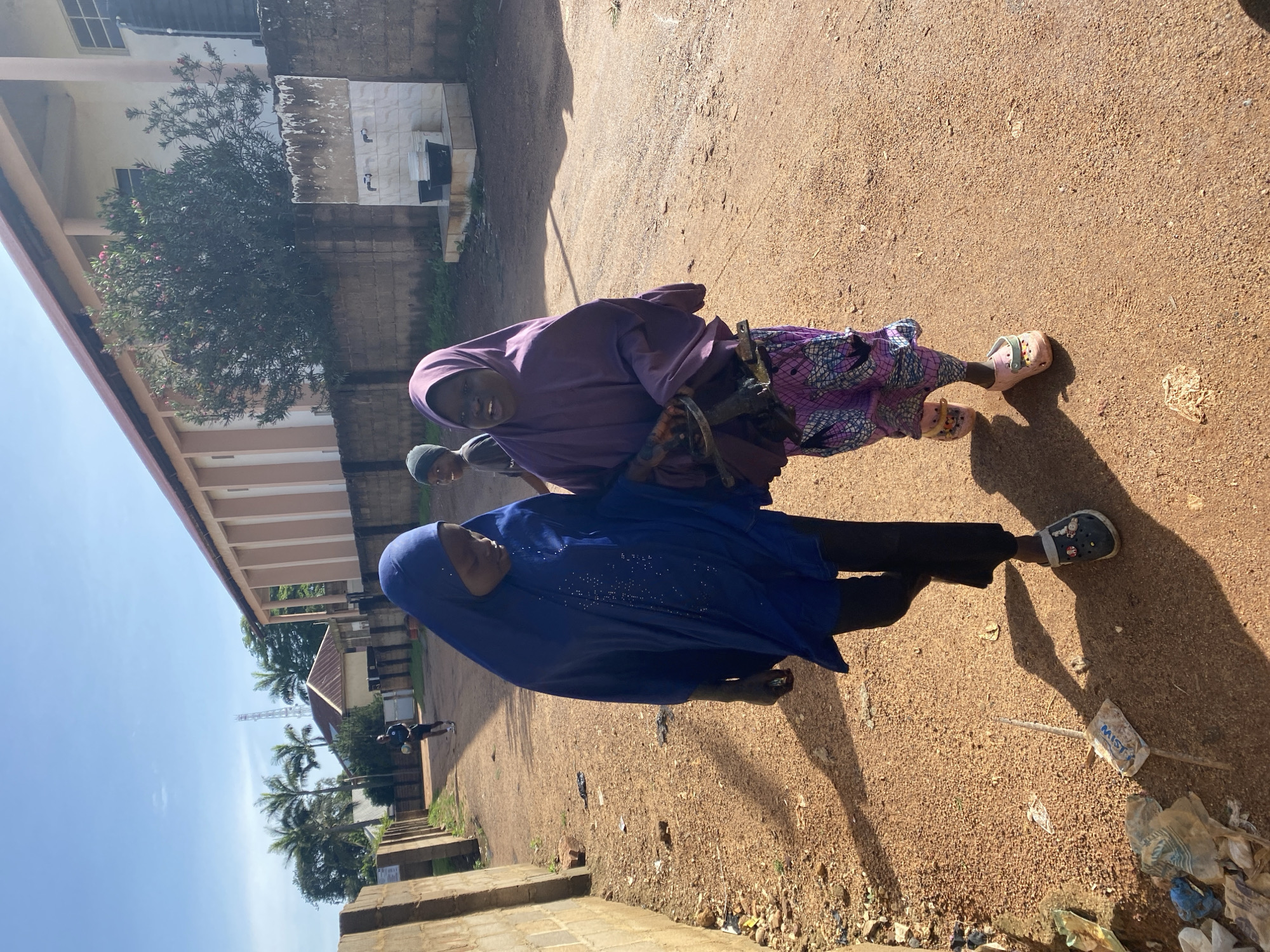 Two young girls are walking down a dirt road in Barakin Lerawi, Nigeria. The girl on the left is wearing a blue hijab and a dark blue dress. The girl on the right is wearing a purple hijab and a colorful patterned skirt. Both girls are wearing sandals. The road is lined with a brick wall on the left and a building with a red roof and a light-colored facade on the right. In the background, there are palm trees and a tall tower. There is some trash on the side of the road. A third, younger child is walking in the background, facing away from the camera.