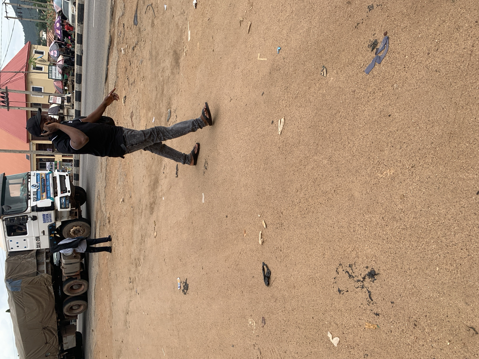 A man in a black polo shirt and distressed jeans is walking and talking on his phone. He is wearing black sandals. He is walking on a dirt road next to a large white truck with a brown tarp covering its cargo. There are several scattered pieces of trash on the ground, including a pair of black sandals and some paper. In the background, there are buildings and a street with cars and umbrellas. This scene appears to be in Adankolo, Nigeria.