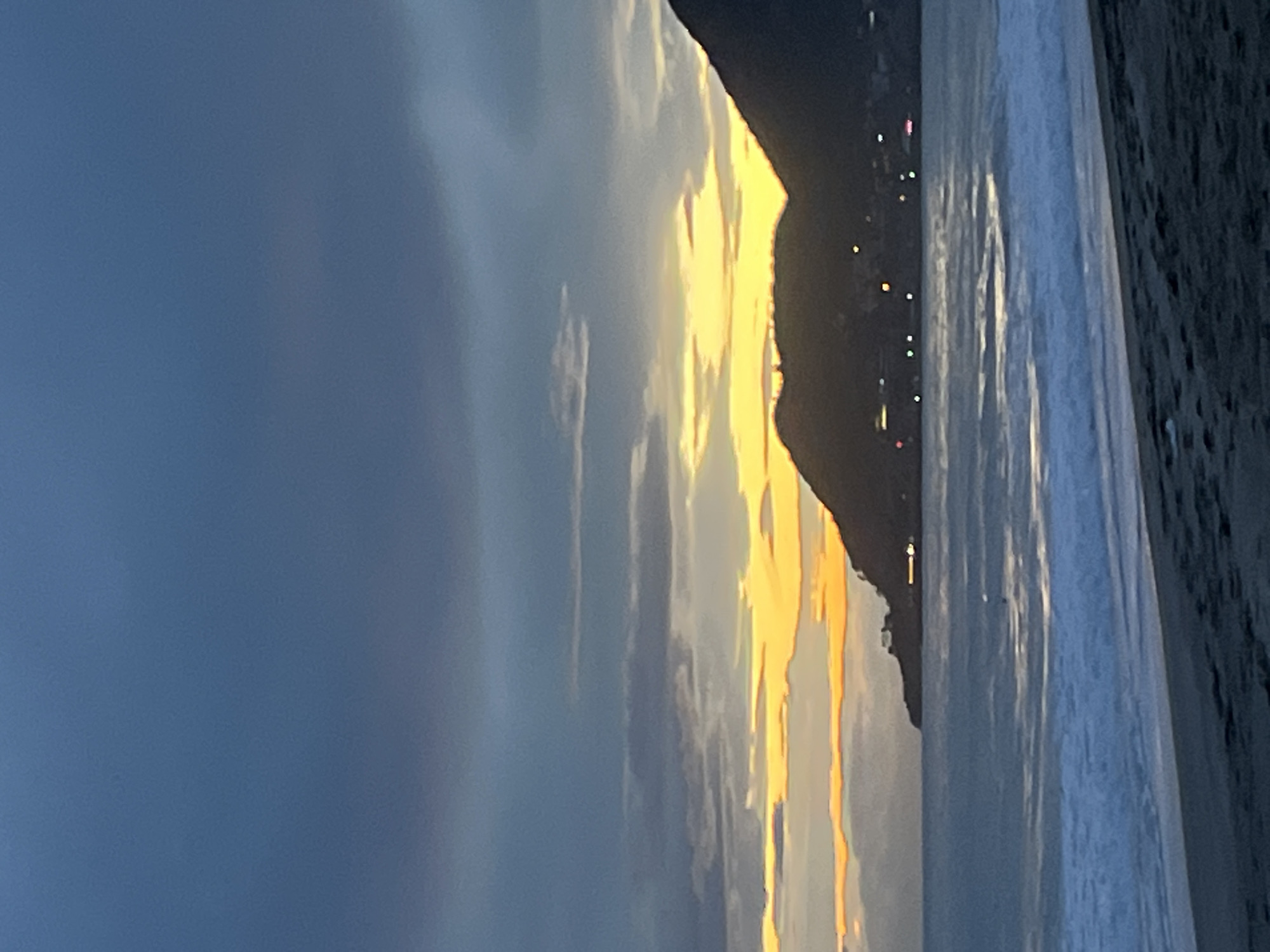 The image captures a seaside scene in Alanya, Turkey. A mountain is silhouetted against the sky, and a string of lights lines the coastline. The sea occupies the right side of the frame, with visible waves. The sky on the left transitions from blue to a vibrant yellow near the horizon. The scene suggests a sunset or sunrise setting.