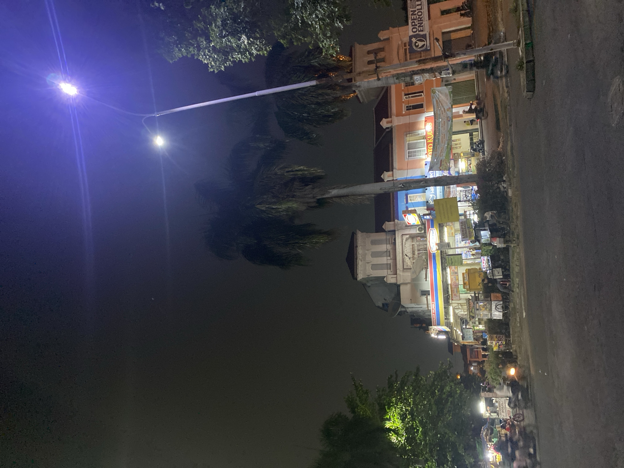 This is a nighttime street scene in Tangerang, Indonesia, likely captured from a low angle. The image depicts a street with several buildings lining the right side, illuminated by streetlights and the lights from within the buildings. The sky is dark, with a bright purple-blue light emanating from a streetlamp in the upper right quadrant, creating lens flares.
The buildings appear to be a mix of commercial establishments, with signage visible, including "Indomaret" and what seems to be an advertisement for "OPEN ENROLLMENT." One building has a light orange facade, while another is blue. In front of the buildings, there are several parked motorcycles and a few people are visible, some walking and others sitting. Two prominent palm trees stand tall in the foreground, their fronds dark against the night sky.
On the left side of the image, a large green tree with bright green foliage is illuminated, possibly by a light source not directly visible. The street itself is paved and appears wet, reflecting some of the ambient light. Several motorcycles are in motion on the street, with their headlights and taillights creating streaks of light due to the long exposure or movement. There are also food carts or small stalls set up along the sidewalk. The overall atmosphere is that of a busy urban street at night.