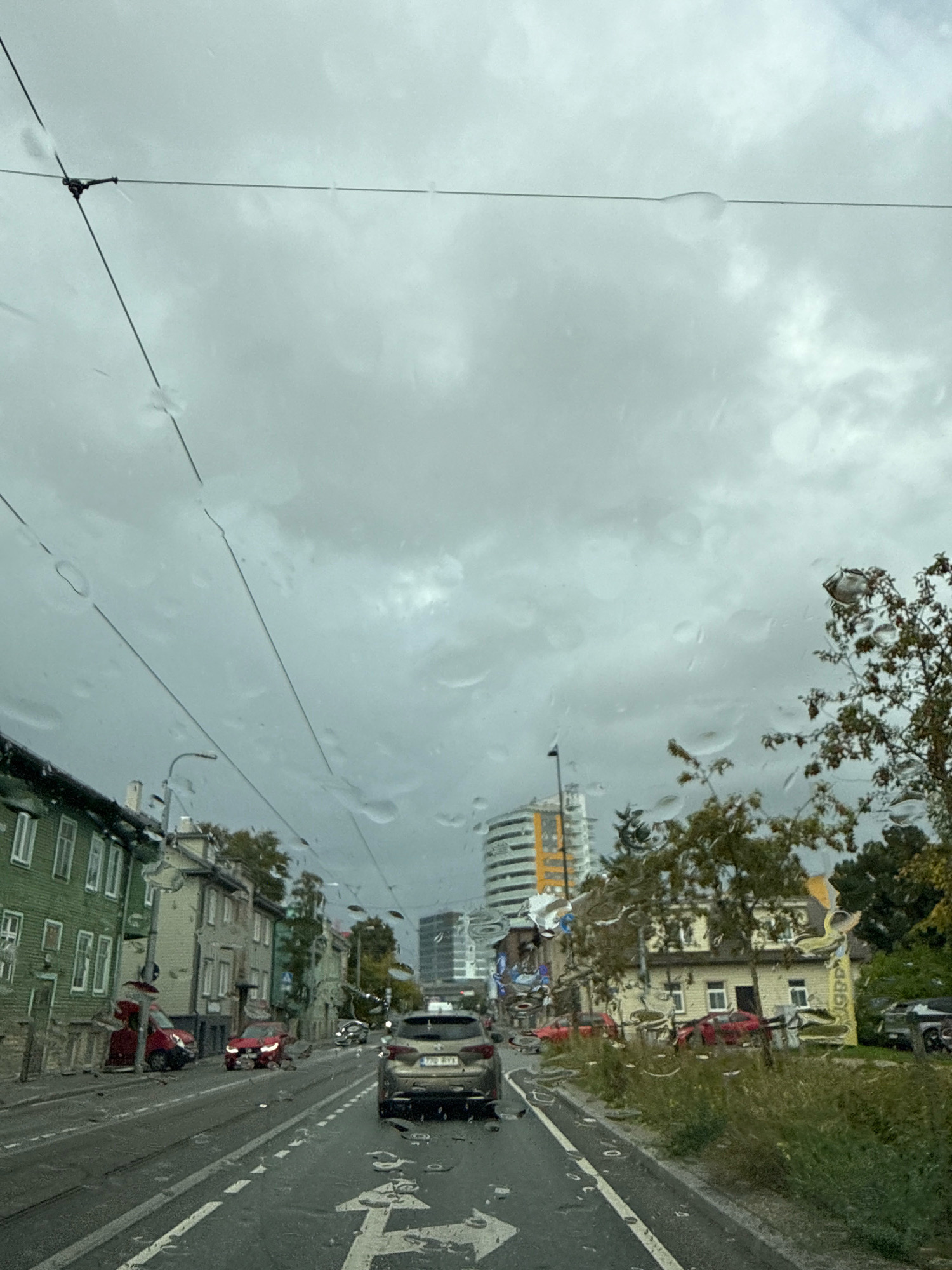 The image shows a street view in Tallinn, Estonia, on a cloudy and rainy day, as evidenced by water droplets on the windshield and the overcast sky. The perspective is from inside a vehicle, looking forward through the front windshield.
In the foreground, the wet asphalt road is visible with lane markings. Several cars are present, including a silver SUV in the right lane with license plate "770 RYX". To the left, a red car is parked, and further down the street, another red car is in motion.
The left side of the street features several traditional wooden houses painted in green and beige. Beyond these houses, modern buildings and construction can be seen, including a tall, cylindrical building with yellow accents. On the right side of the street, more houses are visible, some with vegetation growing in front of them. Trees with autumn foliage are also scattered throughout the scene. Overhead, electrical wires are strung across the street, likely for trams or other public transportation. The overall atmosphere is somewhat subdued due to the weather conditions.
