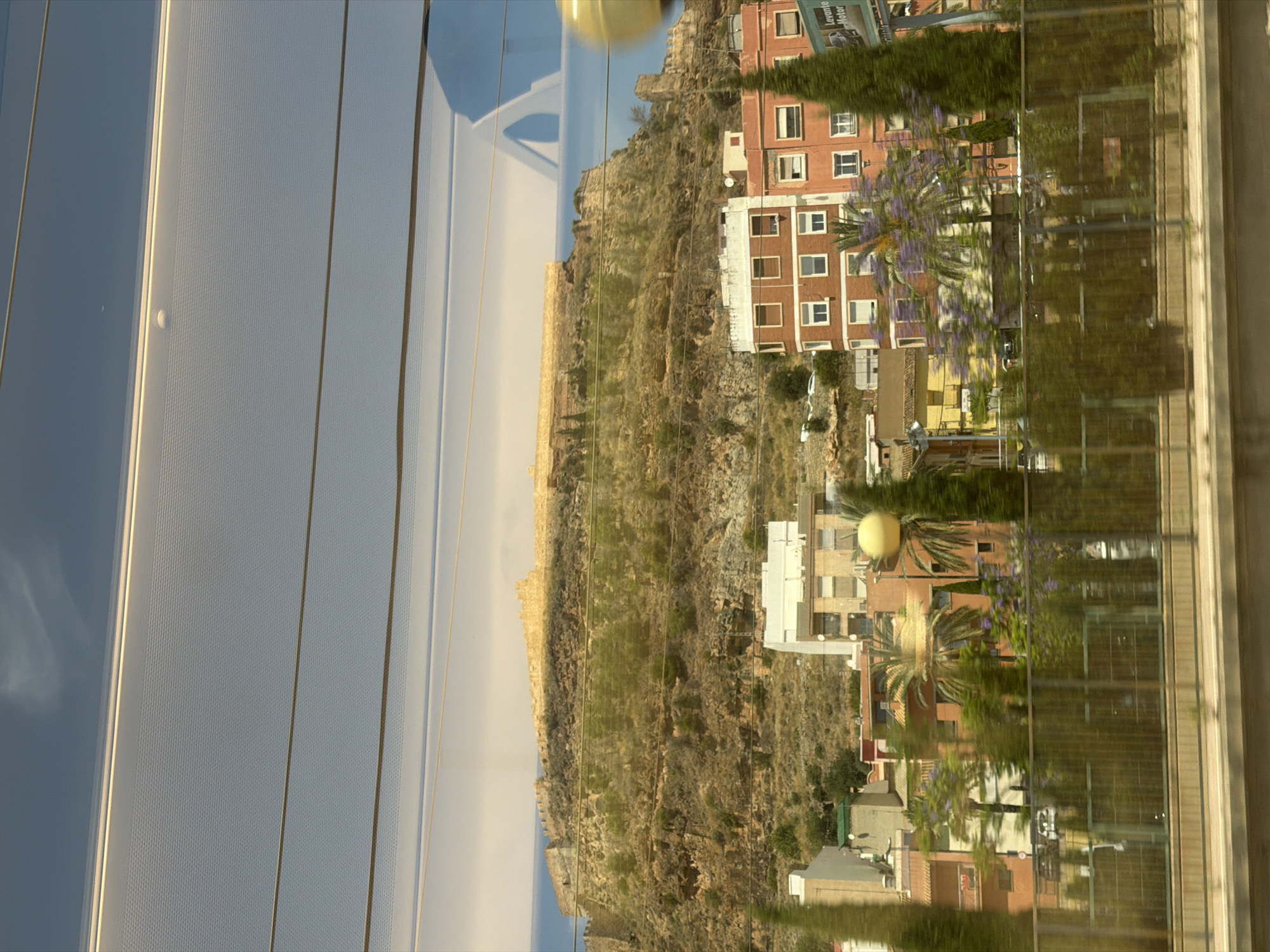The image captures a view of a fortified hilltop overlooking a city, likely taken from a moving vehicle due to the reflections on a glass surface in the foreground. The hilltop features ancient stone fortifications, some sections appearing intact and others in ruins, bathed in sunlight. Below the hill, a cluster of multi-story buildings with reddish-orange and white facades are visible. These buildings appear to be residential or commercial, with visible windows. Green trees, including tall, slender ones and palm trees, are interspersed among the buildings and on the slopes of the hill. A metal fence runs across the lower part of the image, suggesting a separation between the viewer's vantage point and the urban area. The sky above is a pale blue, with subtle cloud formations and prominent horizontal lines, likely reflections from the vehicle's interior or exterior. In the far distance, a sliver of the sea is visible. The scene is characterized by a blend of historical architecture and modern urban development, set against a natural landscape. The reflection of a blue object, possibly a car or structure, is visible on the glass surface in the upper right quadrant. A yellow spherical object, likely a light fixture, is also present in the middle right of the image. The overall impression is of a sunny day in a coastal city with a rich historical past. The area depicted appears to be the Castillo de Peñíscola, a historic fortress located in the city of Peñíscola, although the question specifically mentions Castelló de la Plana. It's possible this is a different location within the province of Castellón, or the provided location is incorrect for the visual elements presented. However, based on the visual evidence, the scene strongly resembles a historic fortified castle atop a hill overlooking a town, a common sight in many Spanish coastal cities.