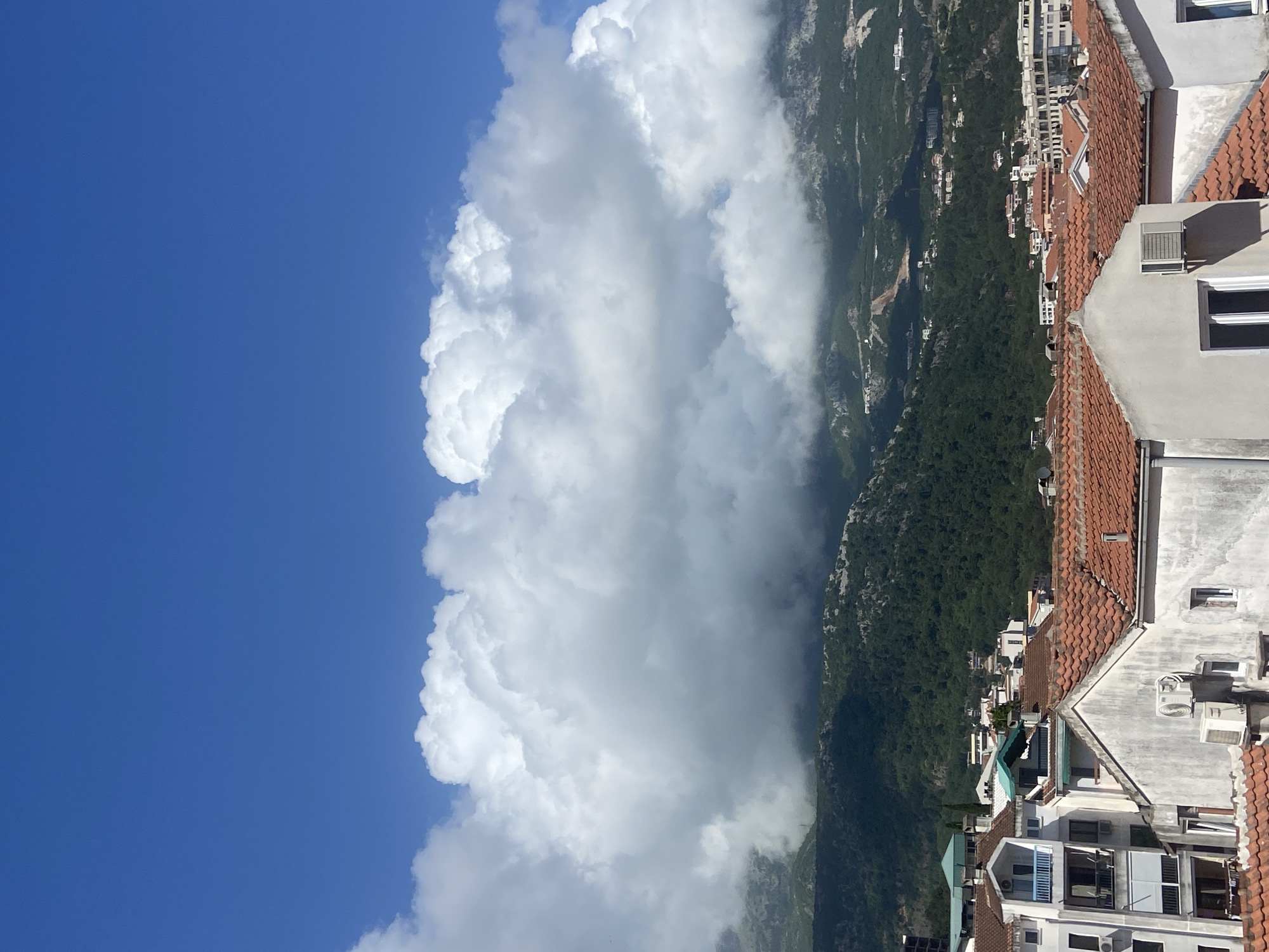 The image shows a view in Budva, Montenegro, with the camera angled upward. In the right foreground, there are buildings with red-tiled roofs and white facades, some equipped with air conditioning units. Behind the buildings, there is a green mountainous landscape. A large, cumulonimbus cloud formation occupies a significant portion of the left side of the image, contrasting with a clear blue sky above and to the left. The sky appears to be the background. The picture is focused on the clouds, as the buildings in the foreground are mostly out of focus. The image is rotated so that the ground is on the right and the sky is on the left.