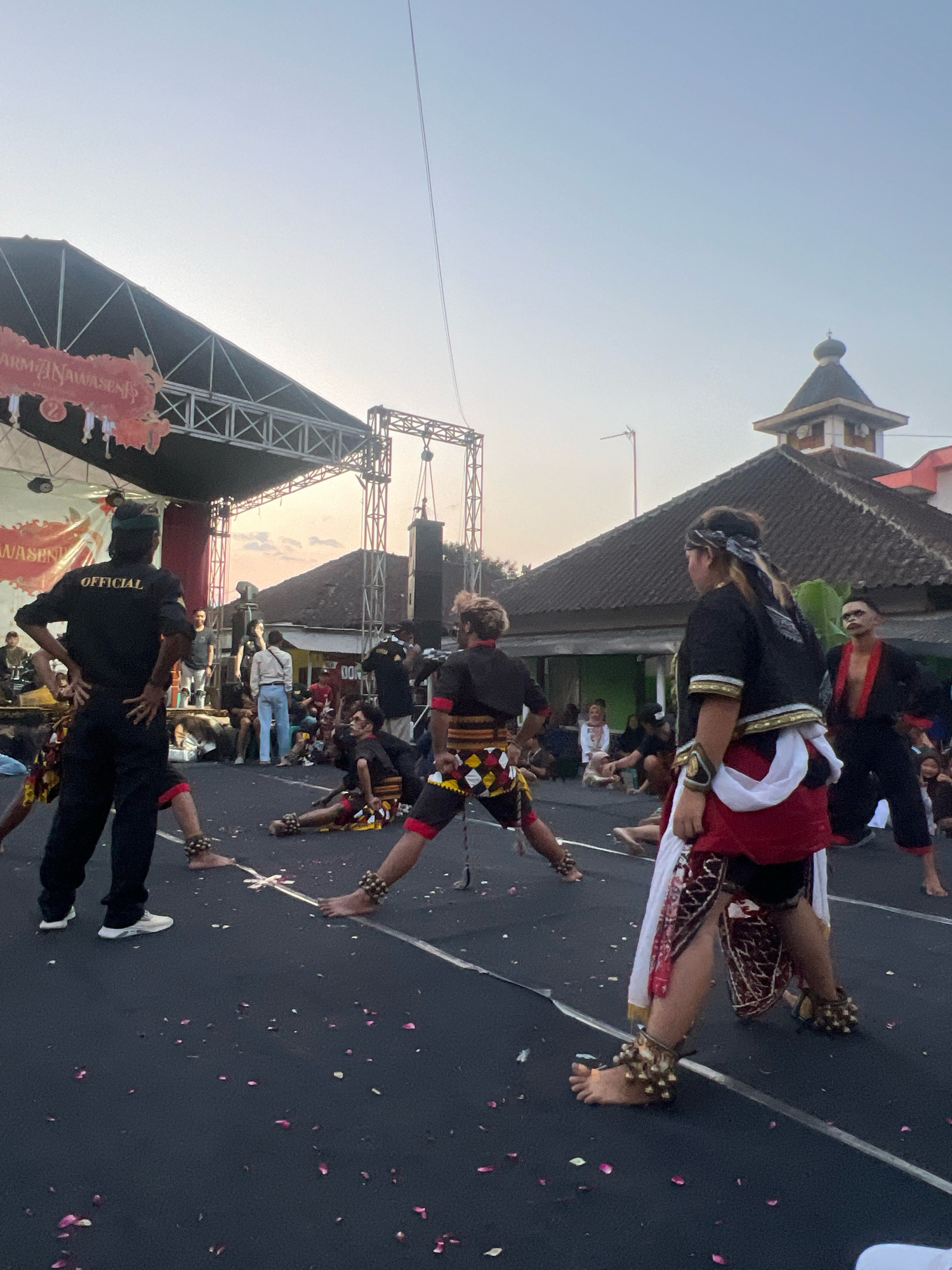 This image depicts an outdoor cultural performance in Madyocondro, Indonesia, likely during the late afternoon or early evening. The foreground features several performers in traditional attire engaged in a dance. One performer, facing away from the camera, wears a black shirt with "OFFICIAL" printed on the back and black pants. Other performers are adorned with patterned loincloths, sashes, and ankle bells. Their poses suggest a dynamic and energetic dance. The ground is dark asphalt, scattered with pink flower petals.
Behind the performers, a stage structure with lighting equipment is visible. Further in the background, buildings with tiled roofs are present, including a structure with a domed roof, characteristic of some religious buildings in Indonesia. A crowd of spectators is seated and standing behind the performance area. The sky is a clear, pale blue with hints of orange from the setting sun. The overall atmosphere suggests a community event or festival.