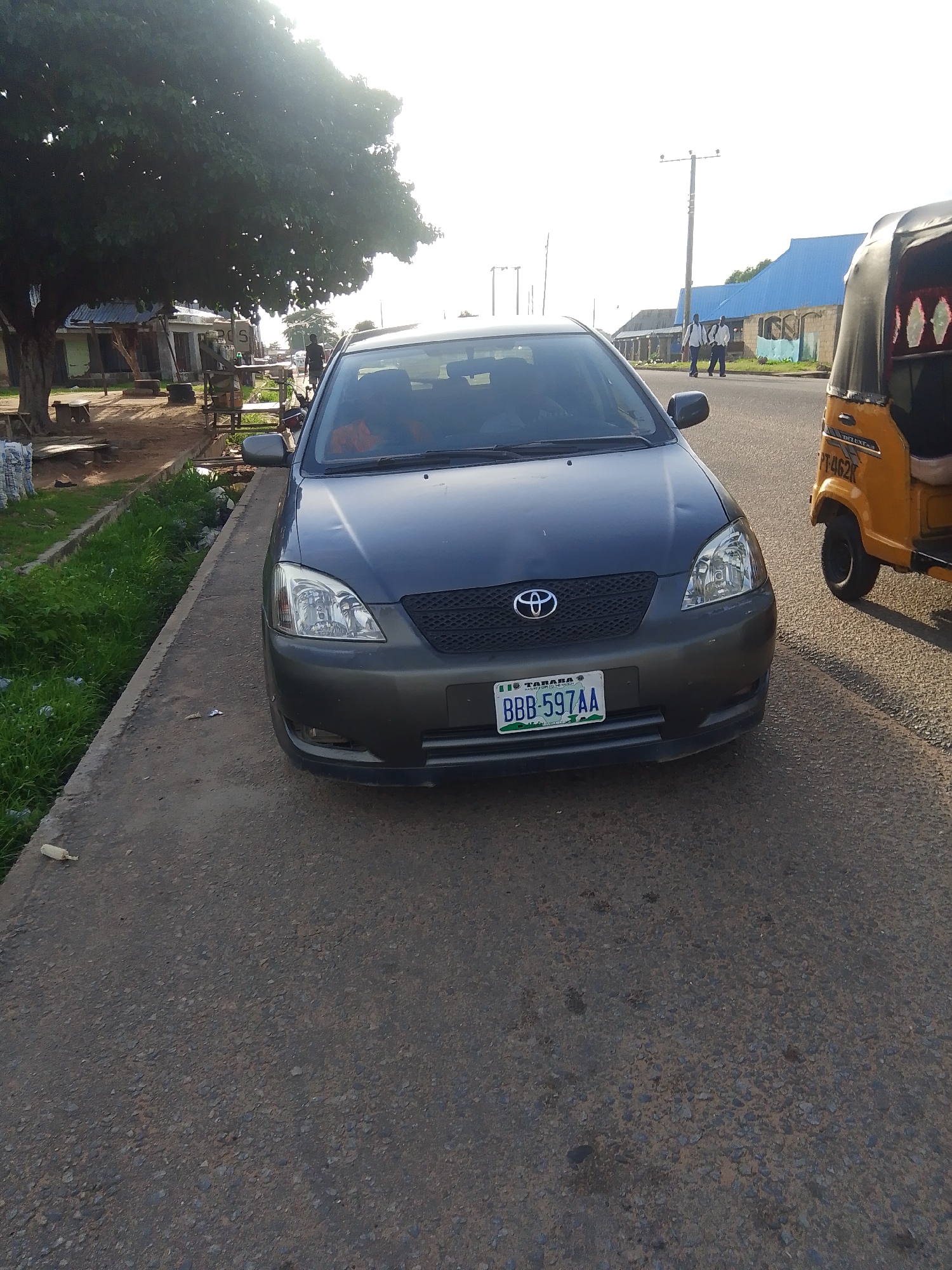This is a frontal medium shot of a dark gray Toyota Corolla parked on an asphalt road. The car's headlights and grill are visible, with a Toyota logo in the center of the grill. A license plate with the text "TARABA BBB-597AA" is attached to the front bumper.
To the right of the car, a yellow auto-rickshaw is partially visible. The auto-rickshaw has "PT-462K" written on its side. In the background, there are buildings with blue and tan roofs, and a tall utility pole. Two young men in white shirts and dark pants are walking on the road in the distance.
To the left of the car, there is a large tree with dense foliage, casting a shadow. A concrete ditch with green grass and some litter runs alongside the road and car. There are also some makeshift stalls and benches under the tree, suggesting a roadside market or waiting area. The sky is bright, indicating daytime, possibly late afternoon due to the angle of the sun and the shadows. The overall scene appears to be a street in a town or city, likely in Nigeria given the location context of Jalingo. The atmosphere seems calm and ordinary, with no obvious emotional cues.