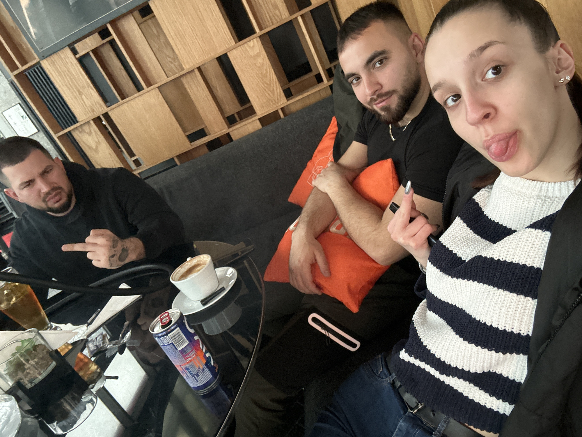 Three people are sitting at a table in what appears to be a café or restaurant. The location may be in Varna, Bulgaria, though this is not explicitly shown. A man on the left is seated at the table, giving the middle finger. Next to him on the table is a partially full drink in a tall glass, a cup of coffee, and a can of Red Bull. A man and a woman are sitting together in a booth. The man is holding an orange pillow, and the woman is sticking her tongue out. The woman is wearing a white and navy striped shirt, and the man is wearing a black shirt. A wooden wall decor is behind the couch.