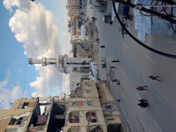 The image captures a wide street in Khan Yunis, Palestinian Territory, under a sky with scattered white clouds. A prominent feature is a heavily damaged mosque in the mid-ground, its minaret standing but visibly fractured and incomplete, and the main structure largely reduced to rubble around its dome. To the left, a multi-story residential building exhibits extensive structural damage, with several floors exposed, revealing debris, twisted metal, and personal belongings. Piles of rubble are distributed throughout the scene, particularly around damaged buildings and along the street's edges.
The paved street, marked with two white lanes, shows varying levels of pedestrian activity. In the foreground, approximately four individuals, including adults and children, walk on the left side of the street, while one individual runs diagonally across the street toward the right. Other small groups of people, including adults and children, are dispersed further down the street and in the background, walking or standing amidst the debris and near the damaged structures. A red sedan is parked on the right side of the street in the mid-ground. In the lower right corner, a white tent structure with Arabic text is partially visible, obscured by dark wires and a pole. Buildings in the background show a mix of intact and damaged structures.