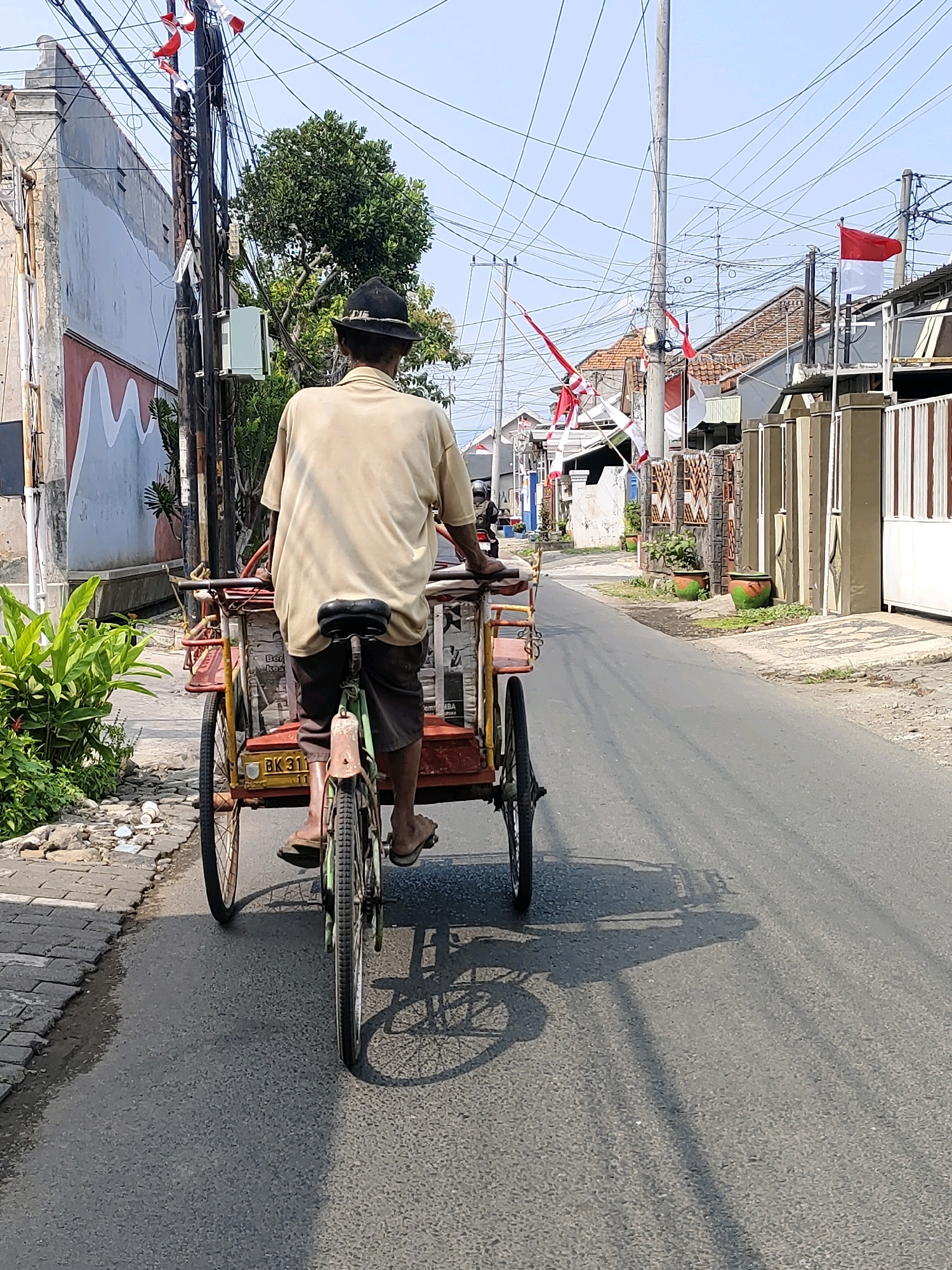 An elderly man wearing a dark hat and a light-colored t-shirt rides a bicycle rickshaw down a paved street in Probolinggo, Indonesia. The rickshaw has a red frame and a yellow cargo box, with a black seat. The man's legs are visible, wearing brown shorts and flip-flops. Indonesian flags, red and white, are visible on poles and strung across the street. The street is lined with houses and utility poles with numerous wires. Lush green plants are on the left side of the street, and shadows are cast on the asphalt. The sky is clear and blue.