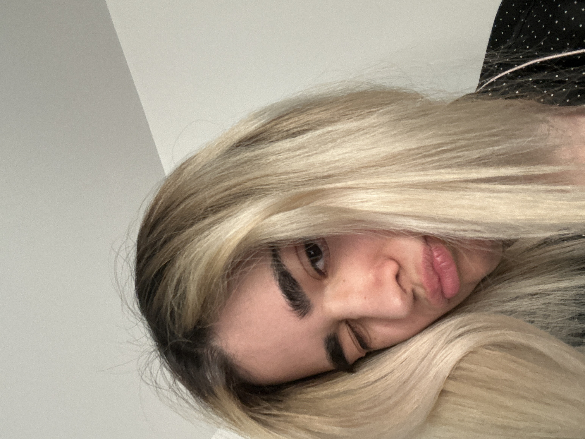 A headshot of a person is presented, taken at an angle. The individual has light blonde hair with darker roots, styled in a straight manner. Their face is positioned to the right side of the frame, featuring one eye open and the other squinted. The person is pouting, drawing attention to their lips. A portion of what appears to be a black pajama top with white polka dots is visible in the upper right corner. The background is a plain, light gray wall. There is no indication that the image was taken in Novate Milanese, Italy.