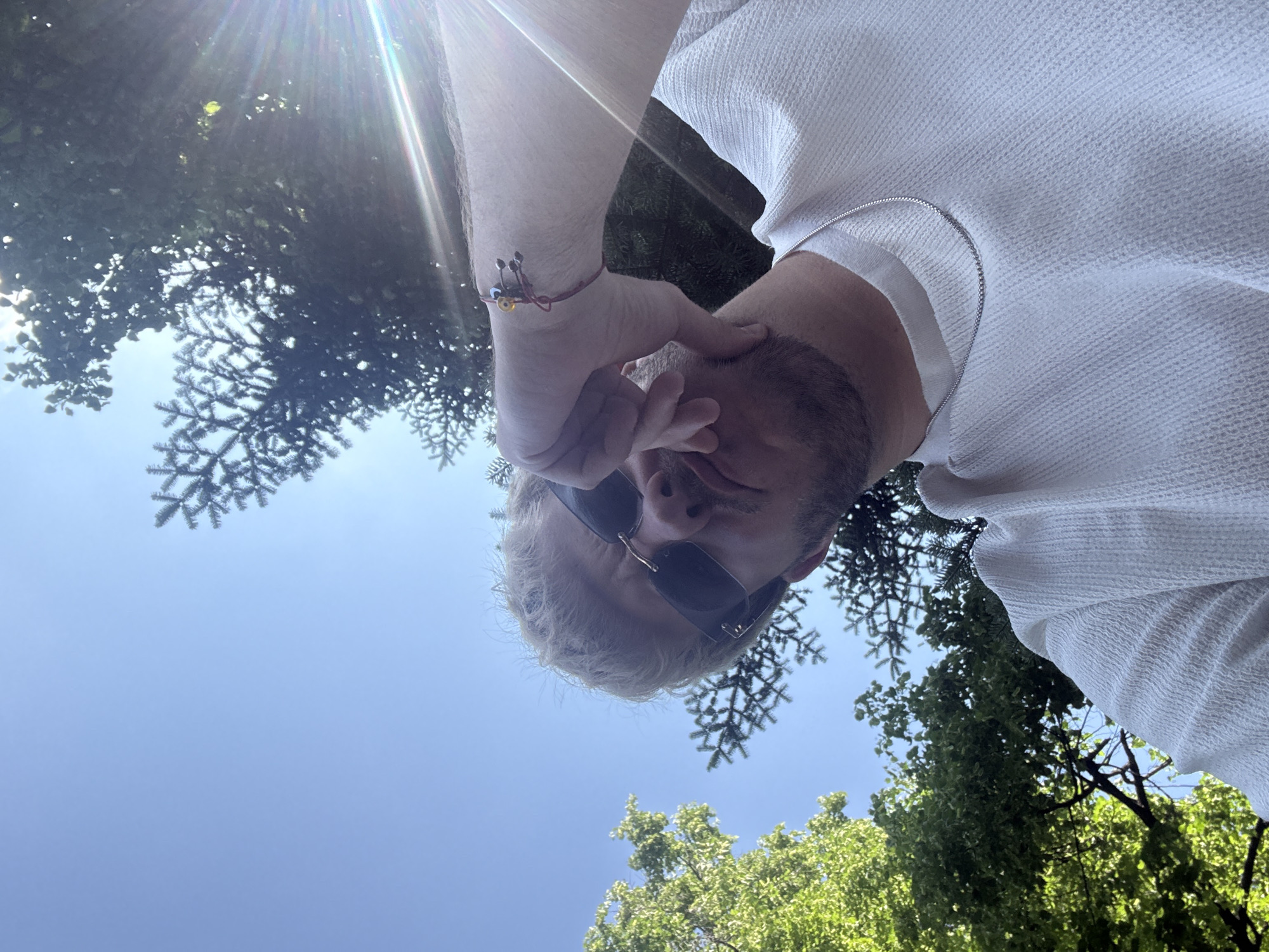 The image captures a unique, low-angle perspective looking upwards, featuring a person seemingly inverted against a bright sky and green trees. The primary subject is a male with light-colored hair and stubble or a short beard, wearing dark sunglasses that obscure his eyes. He is dressed in a white, textured shirt, under which a thin silver chain is visible. On his right wrist, he wears a colorful bracelet adorned with small beads and a prominent evil eye charm. One of his hands is positioned near his face.
The background consists of a clear, bright blue sky with significant sun flares emanating from the upper left, indicating a sunny day. Lush green trees frame the scene, with distinct needle-leafed branches visible, suggesting the presence of conifers, alongside broad-leafed trees. The setting appears to be an outdoor natural environment, likely a park or garden, consistent with locations found in Sofia, Bulgaria. The overall impression is a casual outdoor portrait or selfie taken from an unconventional viewpoint. No text is visible in the image.