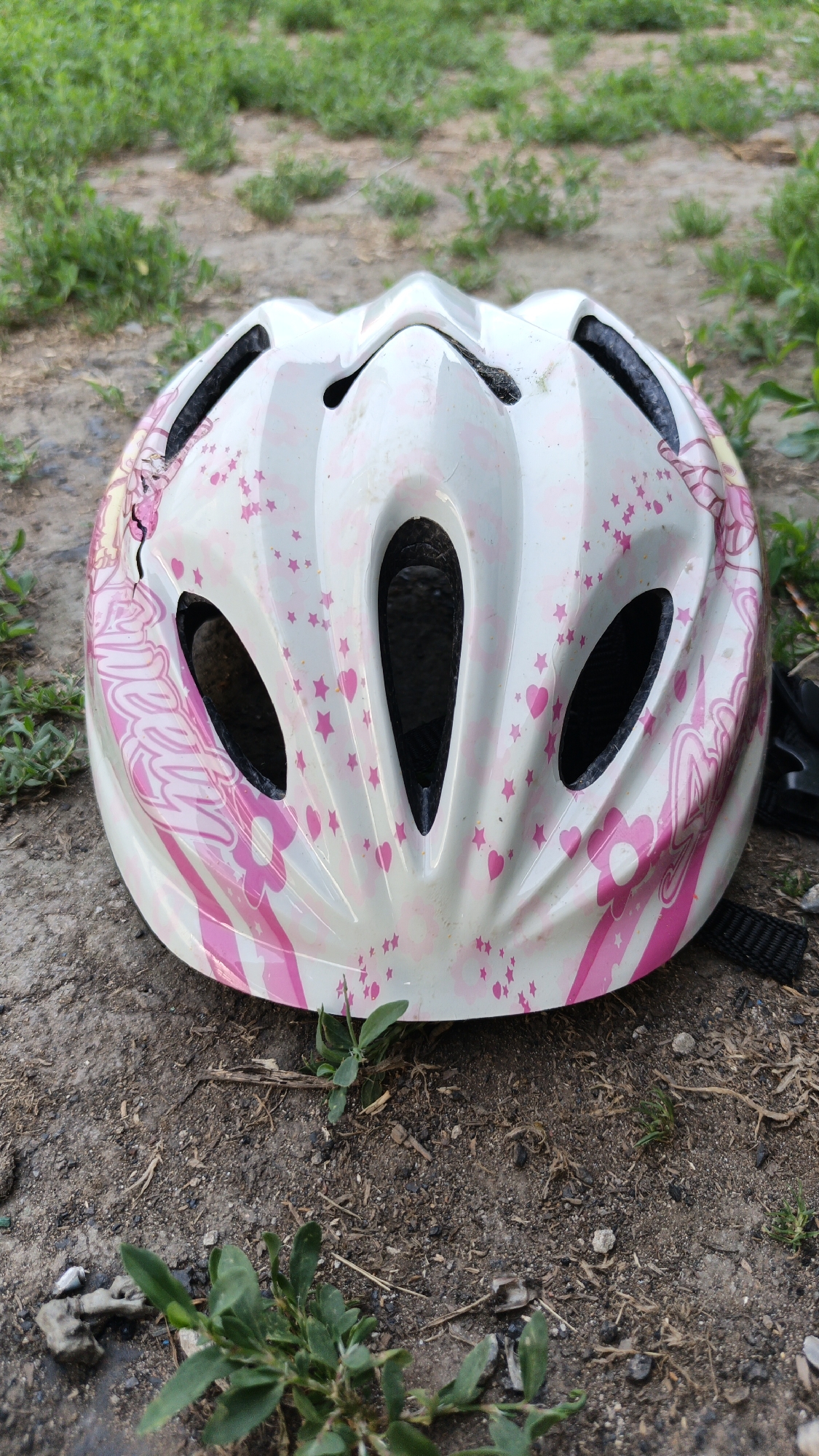A white bicycle helmet with pink stars and flowers is lying on the ground. The helmet is tilted forward and slightly to the left. The ground is covered in dirt, small stones, and green grass. The background shows more green grass. There are no people visible in the image.