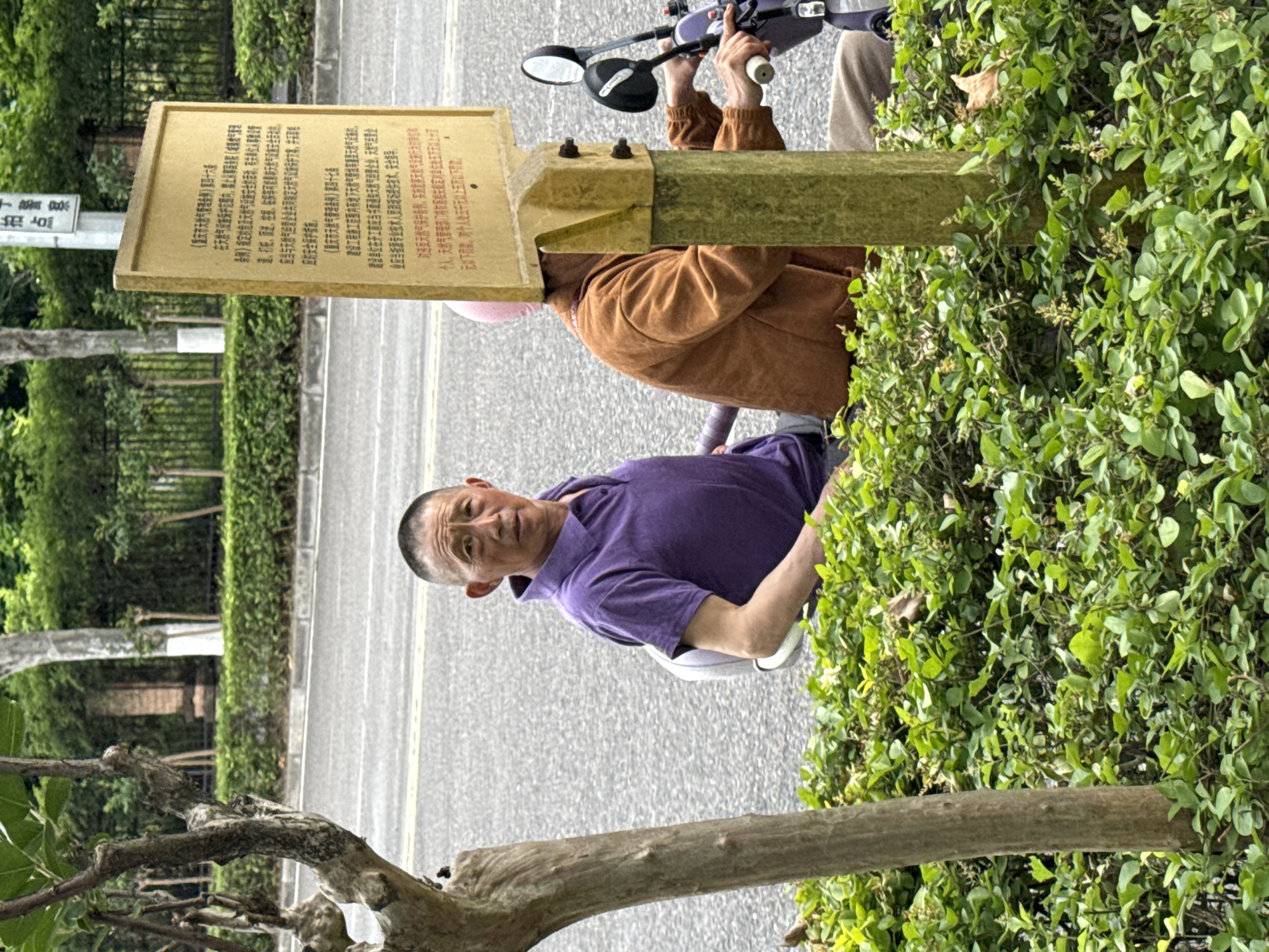 The image depicts a man in a purple shirt seated on a scooter. He is positioned next to a sign with Chinese text. Another person in a brown jacket is visible on the scooter as well. The sign is mounted on a wooden post surrounded by greenery. In the background, there is a road and additional foliage. The OCR text on the sign mentions "重庆市" (Chongqing City) and "天然气" (natural gas), suggesting the location is in Chongqing. The signs point towards this image being taken in Jiangjin, China.