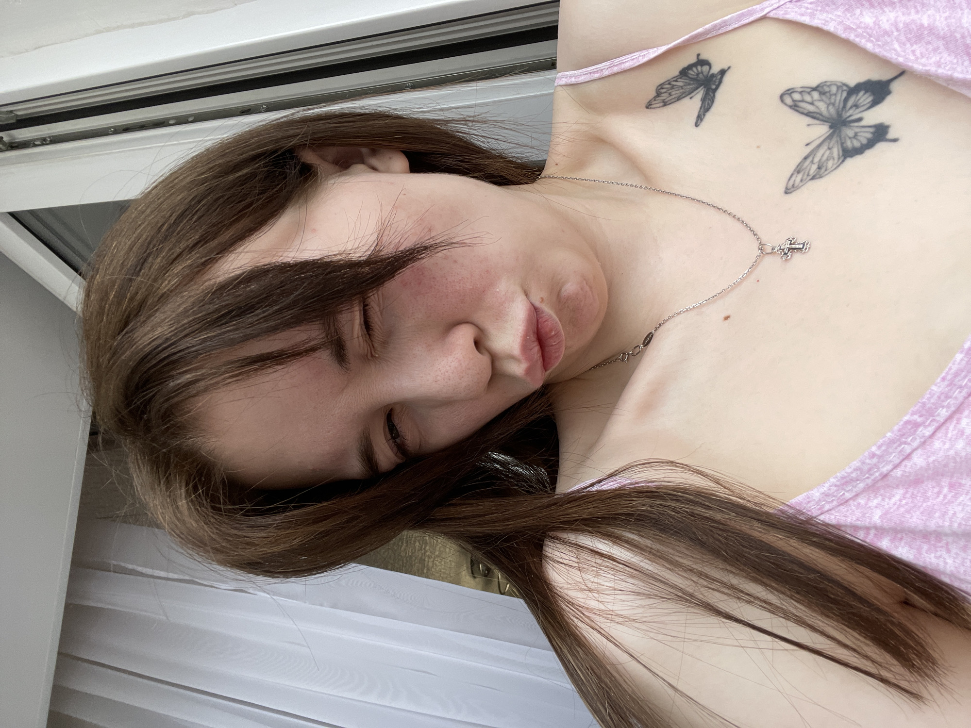 A young woman with fair skin and long, brown hair is lying down. She is wearing a pink tank top, and a silver necklace with a cross pendant is visible around her neck. She has two butterfly tattoos on her upper chest. The background suggests a window with white curtains. The city of Chaikovsky, Russia is not discernable from this image.