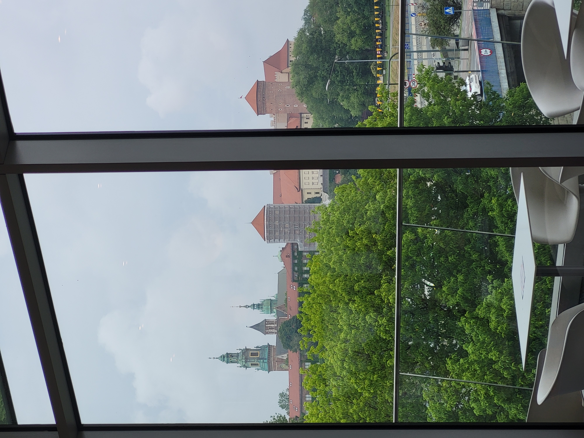 The image presents a daytime view from inside a modern building, looking out through large, dark-framed windows onto an urban landscape under an overcast sky. In the foreground, parts of white, modern chairs and tables are visible inside.
The outdoor scene is dominated by a historical architectural complex, identifiable as Wawel Castle and Cathedral, situated on a tree-covered hill. The castle features multiple towers with reddish-brown roofs and walls, some showing scaffolding, suggesting ongoing maintenance or construction. Below the main castle, the Wawel Cathedral's distinctive green spires, domes, and turrets are clearly visible amidst more trees and buildings.
At the base of the hill, a road with a white car and a bridge are visible. Streetlights line the road, and yellow triangular banner flags are strung along a railing. Several European road signs are present, including a pedestrian crossing sign, a directional arrow, a circular speed limit sign displaying "30", and a circular height restriction sign indicating "3m". The landscape is rich with vibrant green trees. The sky is uniformly light gray, indicating an overcast day. The scene strongly suggests a view of Wawel Castle in Kraków, Poland, likely from across the Vistula River.