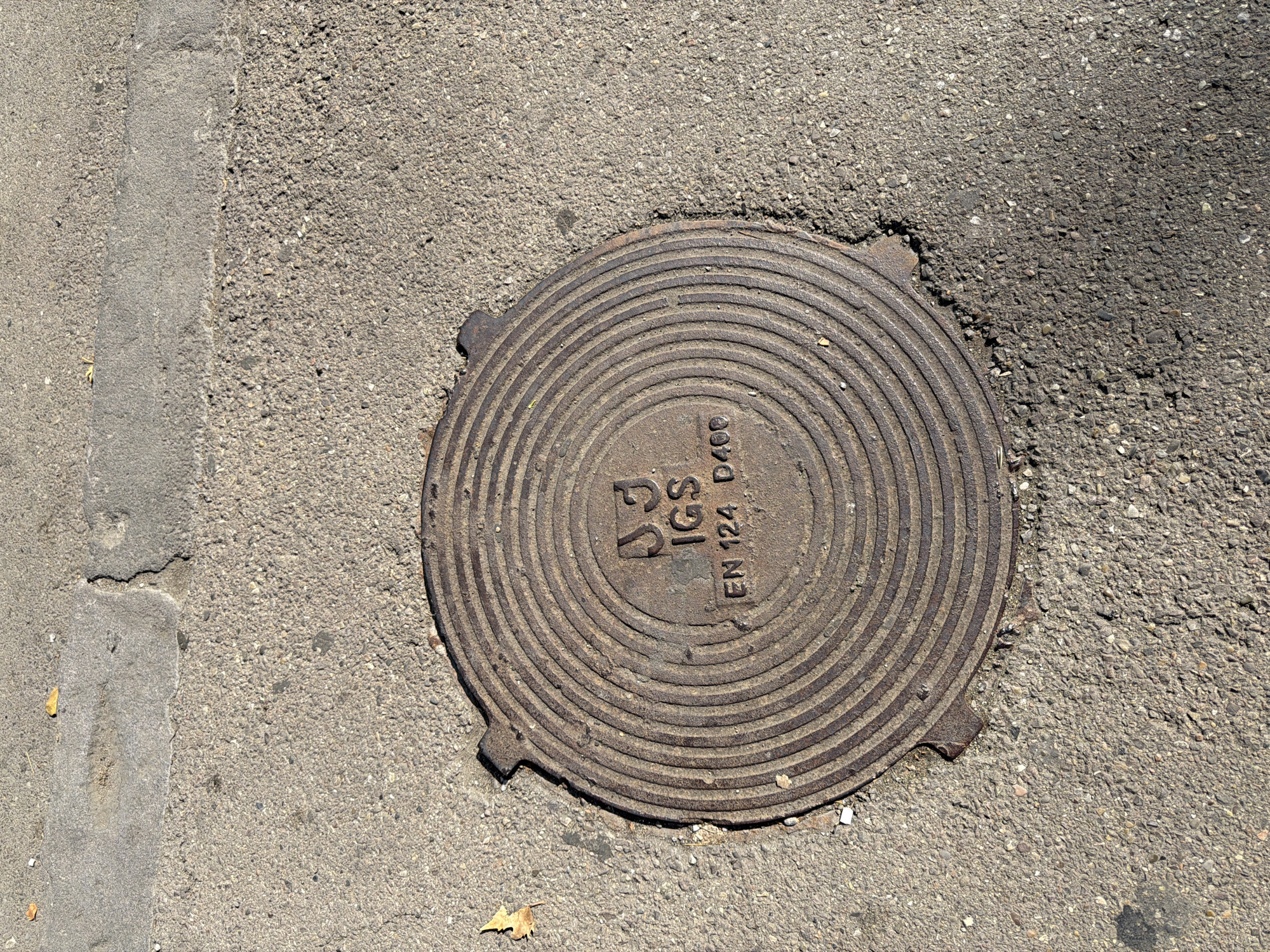 A circular, cast-iron manhole cover is centered on a paved surface in Tbilisi, Georgia. The cover features concentric raised rings and a raised inscription in the center. The inscription reads "JJ IGS EN 124 D400". The surrounding pavement is a mix of asphalt and concrete, with some small debris and a fallen leaf visible. The lighting suggests it is daytime, with a shadow cast by an unseen object to the right.