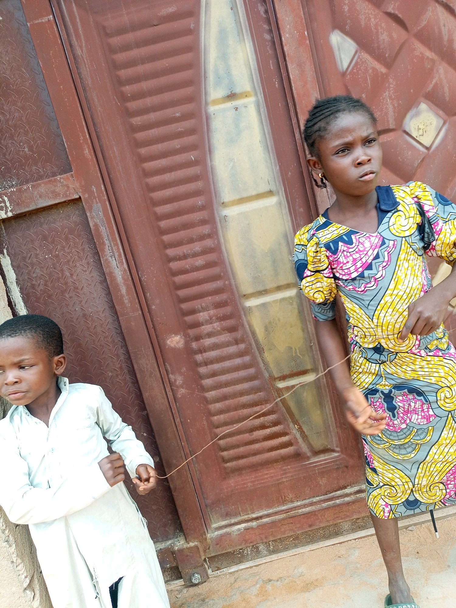 Two children are standing in front of a weathered, brown metal door in what appears to be an outdoor urban setting. The child on the left is a young boy wearing a light-colored, long-sleeved shirt and pants. His left hand is holding a thin, brown string that extends towards the child on the right. The child on the right is a young girl wearing a colorful dress with a yellow, blue, and pink pattern. She is also holding the same string in her right hand, and her body is turned away from the camera, looking to the right. The door has a textured surface with vertical ribbed sections and diamond-shaped patterns. The ground in front of the door is dusty and uneven. The overall scene suggests a casual, everyday moment in a Nigerian city like Tempure.