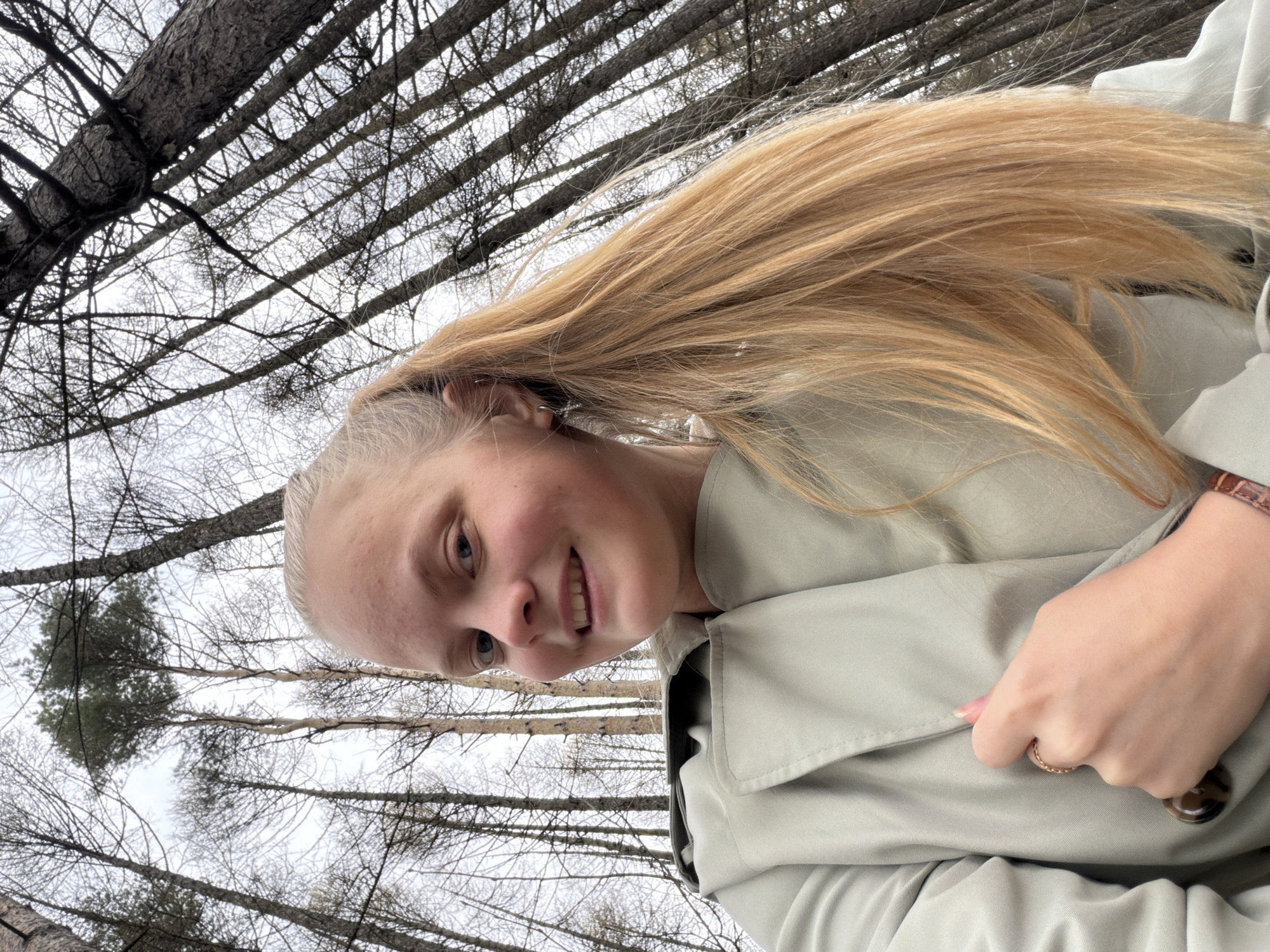 A young woman with long, light blonde hair is posing for a selfie in a forest. She is wearing a light green trench coat and is holding the button of the coat. The background consists of tall, bare trees with a cloudy sky visible through the branches. While the precise location cannot be determined from the image alone, the request specifies Izhevsk, Russia. Therefore, the image depicts a woman posing in a forest in or near Izhevsk, Russia.