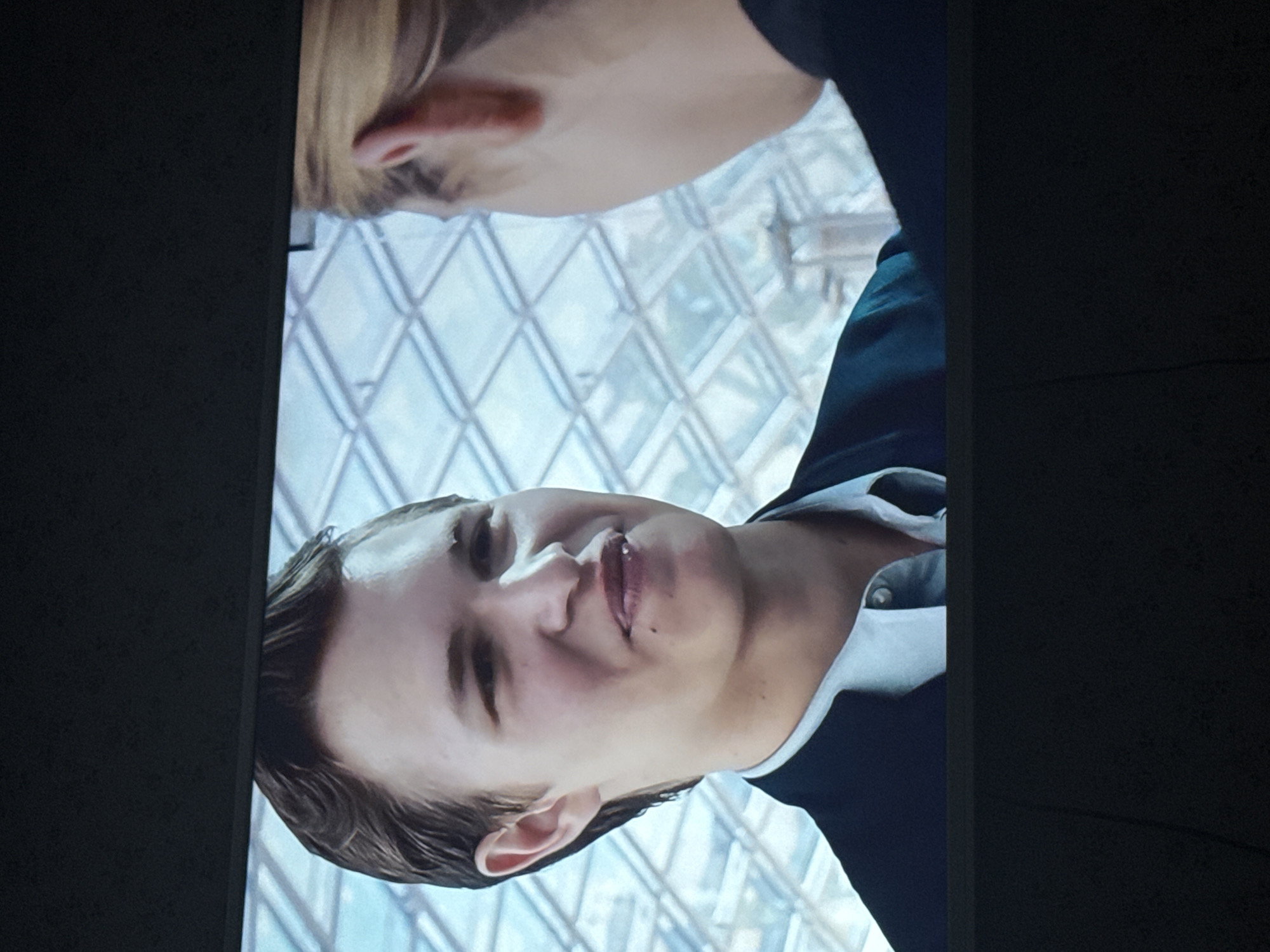 A medium shot depicts a fair-skinned man in a dark suit and white shirt, smiling slightly. He has short brown hair styled to the side. Behind him is an architectural structure with a geometric pattern of glass or light-colored panels, possibly a roof or atrium. The image is tilted slightly clockwise. There is no indication of the location being in Mytishchi, Russia.