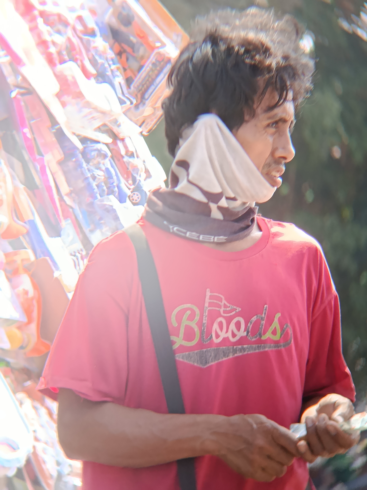 A medium shot captures a man in Desa Kebon Manis, Kecamatan Cilacap Utara, Indonesia, looking to his right with a slightly open mouth. He wears a red t-shirt with the word "Bloods" and two flags printed in green and yellow. A light-colored fabric is wrapped around his neck. He holds a small wad of cash in his right hand, which is in focus. Behind him, a collection of colorful items, possibly merchandise, is visible and blurred. The background consists of out-of-focus greenery. The lighting suggests an outdoor environment, possibly a street or market.