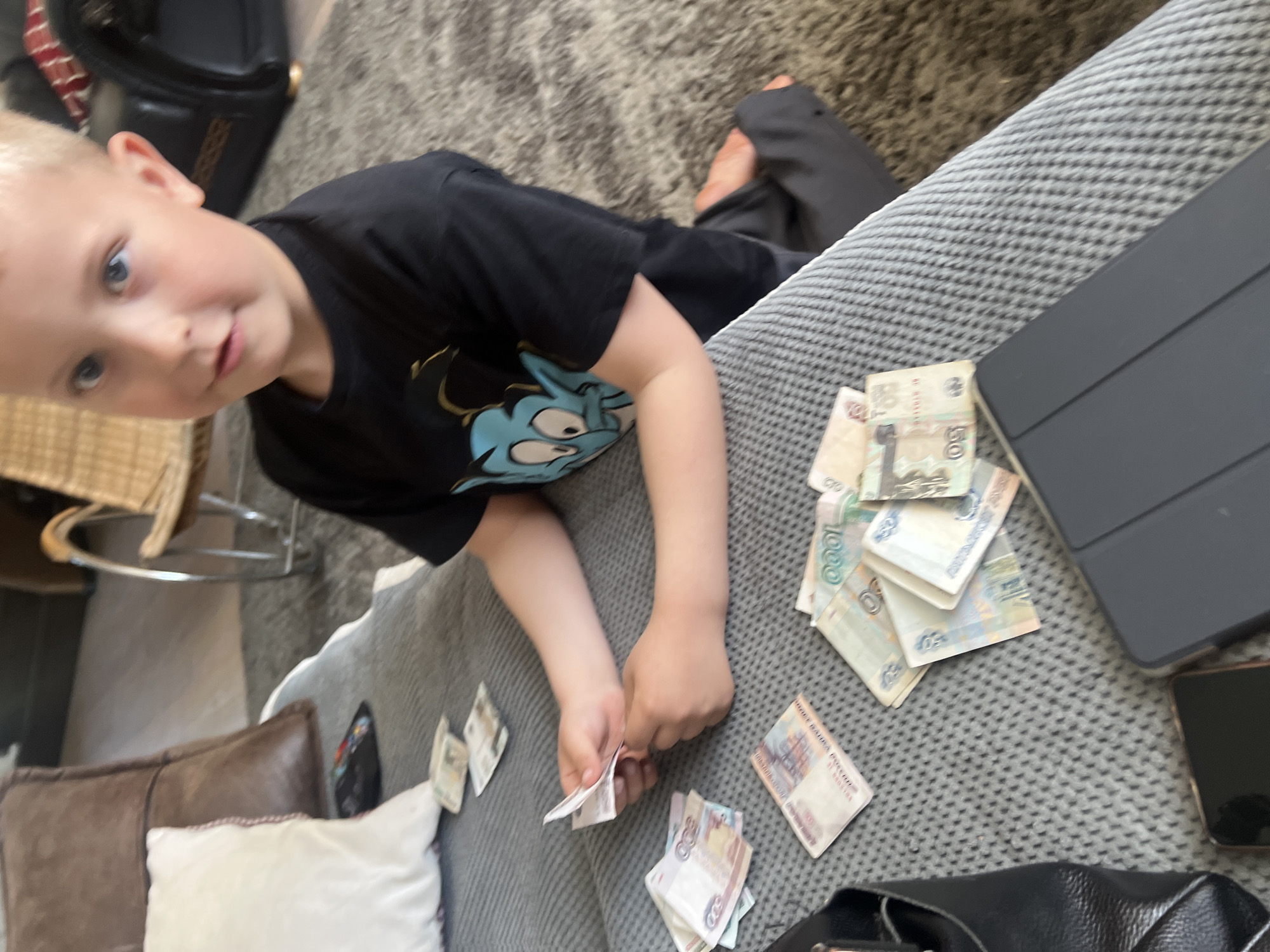 A young boy with blond hair and blue eyes is lying on a gray, textured sofa. He is wearing a black t-shirt with a blue cartoon graphic. He is looking at the camera while holding several banknotes in his hands. Spread out on the sofa around him are numerous other banknotes, a black tablet, and a black smartphone. In the background, there is a gray rug, a chair with a wicker seat, and a brown leather pillow with a white decorative pillow next to it. A black bag is also visible.