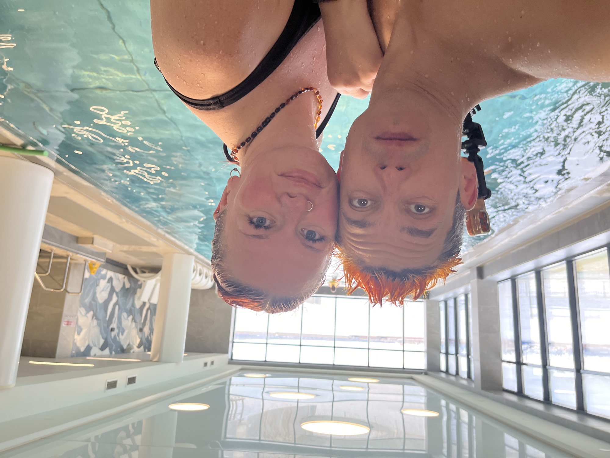 This vibrant image features two individuals, a woman and a man, captured in an unusual, inverted perspective from slightly below the water's surface in an indoor swimming pool in Chaikovsky, Russia. Their faces and upper bodies are visible, covered in water droplets, suggesting they are either emerging from the water or submerged just beneath the surface, looking up at the camera.
The woman is on the left, wearing a black bikini top and an amber-colored beaded necklace. She has blue eyes, a small septum piercing, and other small earrings. Her light brown hair is wet. The man is on the right, with darker hair and strikingly bright orange, spiked hair on top. He has dark eyes. A black device, possibly an action camera, is attached near his right ear. Both individuals are looking directly at the camera with neutral to slightly smiling expressions.
The setting is a modern, brightly lit indoor swimming pool. The clear, blue-tinted water fills the lower portion of the frame, with ripples and reflections visible. The reflections on the water's surface clearly show the pool's architecture, appearing upside down: large floor-to-ceiling windows, white pillars, and circular ceiling lights reflected as bright discs on the water. In the background, there's a wall with an abstract grey and white pattern, below which an inverted white sign with unclear red text can be faintly discerned. The overall impression is one of a playful moment captured in a clean, contemporary aquatic environment. The time of day appears to be daytime, given the natural light filtering through the large windows.