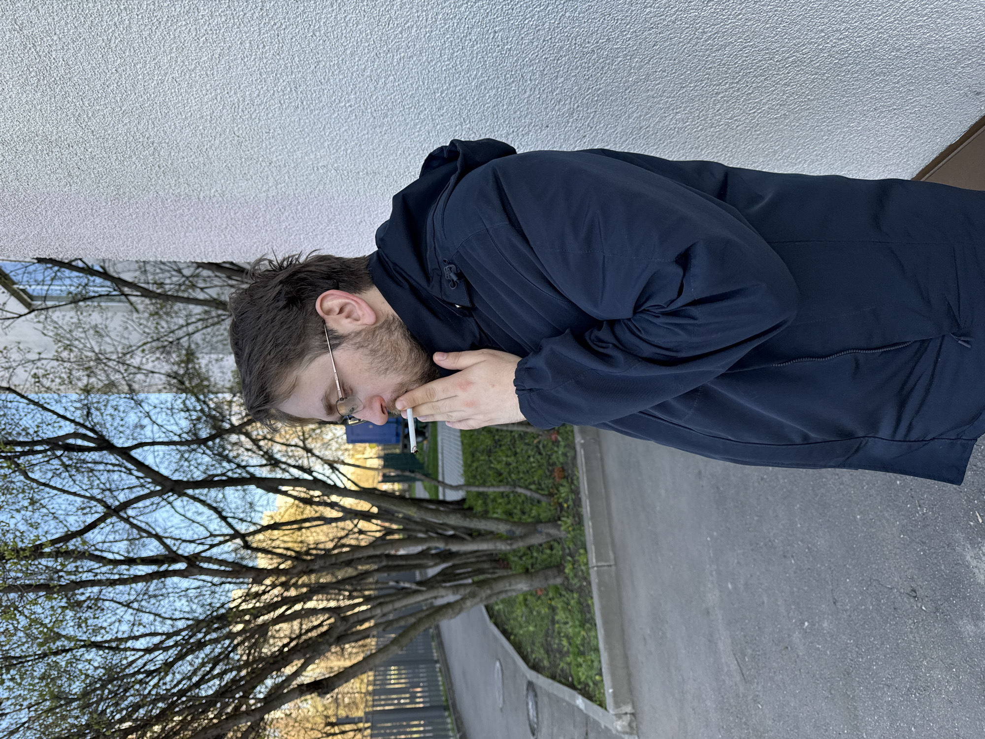 A man with short brown hair, glasses, and a beard is smoking a cigarette outdoors. He is wearing a dark blue hooded jacket. He appears to be standing on a paved surface near a bare tree and some grass. In the background, there is a light-colored building. While Moscow, Russia, cannot be explicitly verified, there is no basis to presume otherwise.