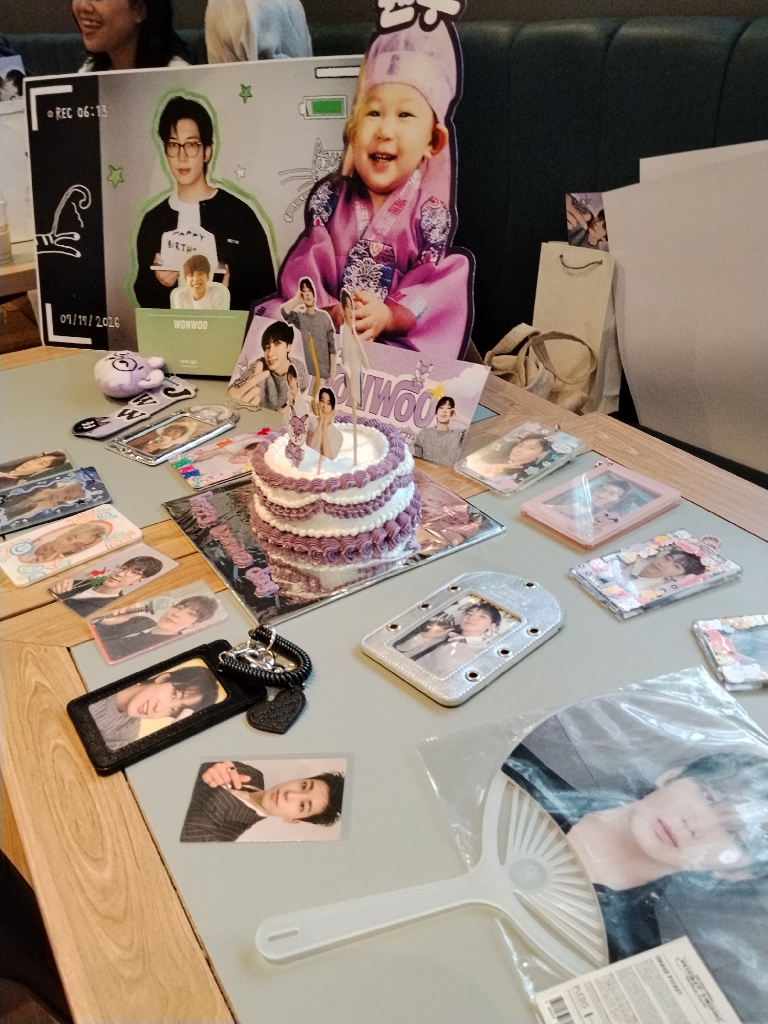The image depicts a table decorated with K-pop merchandise, specifically for an artist named "WONWOO." The setting appears to be a casual gathering or fan event.
Dominating the background is a large cardboard cutout featuring two images of WONWOO. The top image shows him wearing glasses and holding a sign that says "HAPPY BIRTHDAY." Below him is a smaller cutout of a child in traditional Korean attire, smiling. The cutout also has a "REC 06:13" graphic and a date "09/19/2026."
In the foreground, the table is laden with various items:
* A two-tiered cake with purple frosting.
* Numerous photocards and small framed photos of WONWOO.
* Several clear plastic photo holders with pictures of the artist.
* A keychain with a plush toy and a heart-shaped charm.
* A hand fan with a large image of WONWOO.
* A gift bag is partially visible in the background.
The lighting suggests an indoor setting, possibly a cafe or a private room, with the overall atmosphere focused on celebrating WONWOO. The presence of the "09/19/2026" date on the backdrop might indicate a future event or a symbolic date.
There are no explicit details in the image that pinpoint the location to Serang, Indonesia. The provided information about the city of Serang, Indonesia, cannot be confirmed or refuted based on the visual content of the image.