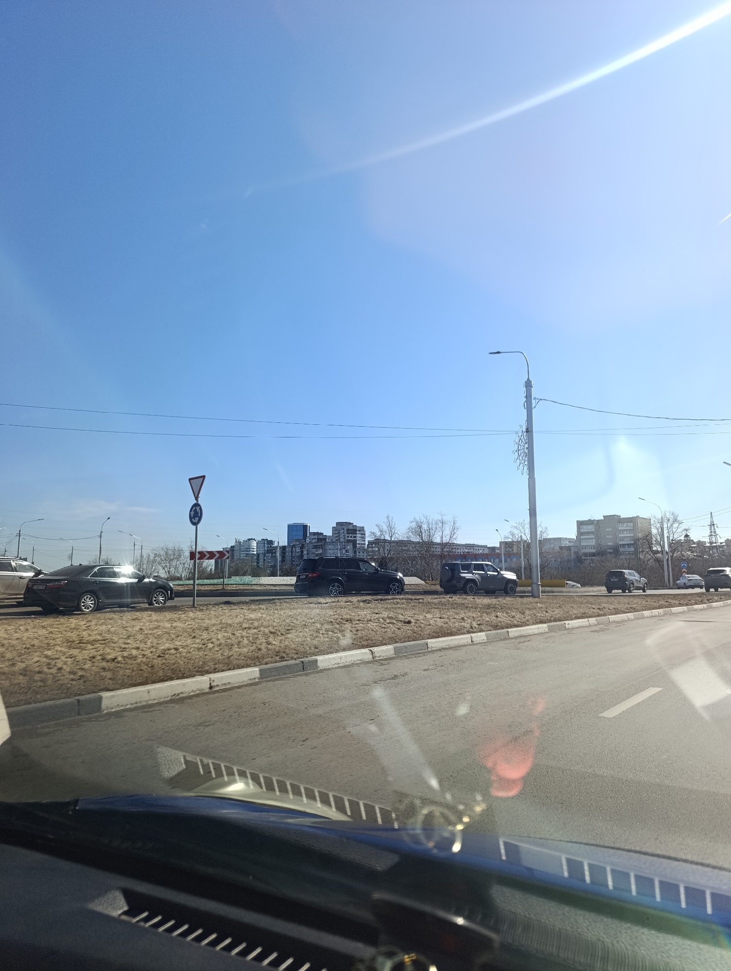 The image depicts a street scene in Krasnoyarsk, Russia on a clear, sunny day. The view is from inside a vehicle, likely the driver's perspective. On the other side of a median strip, several cars are visible, including a dark sedan, a dark SUV, and other vehicles farther down the road. A road sign with a circular arrow indicating direction and a yield sign is present. Beyond the road and the vehicles, there are buildings in the distance, suggesting an urban setting. Street lights line the road. The visible portion of the car's dashboard suggests that the picture was taken from inside the vehicle itself.