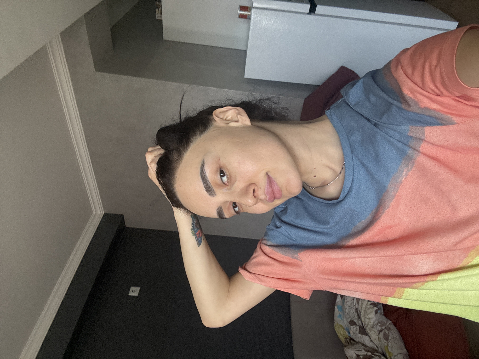 This indoor portrait features a person, likely a woman, with dark hair tied back, looking directly at the camera with a neutral to slightly playful expression. She is wearing a multi-colored t-shirt with prominent sections of blue, pink, and yellow. A thin silver chain is visible around her neck. On her right forearm, a distinct blue and orange butterfly tattoo is present. Her right hand is raised, with fingers resting on the top of her head.
The background suggests a modern interior setting, possibly a room in an apartment. On the left side of the frame, there's a light grey wall with white decorative molding along the top edge, leading to a darker grey section. Further back and above, white cabinetry or furniture, possibly kitchen elements, is visible. On the right side, a dark, possibly black, door or wall panel is seen, featuring a small white square object that resembles a light switch or outlet. Below the person's arm, a patterned fabric, potentially a blanket or throw, is visible on what appears to be a bed or sofa. The lighting is consistent with an indoor environment. No visible text is present in the image, and there are no specific visual cues within the image that directly indicate the location as Tbilisi, Georgia.