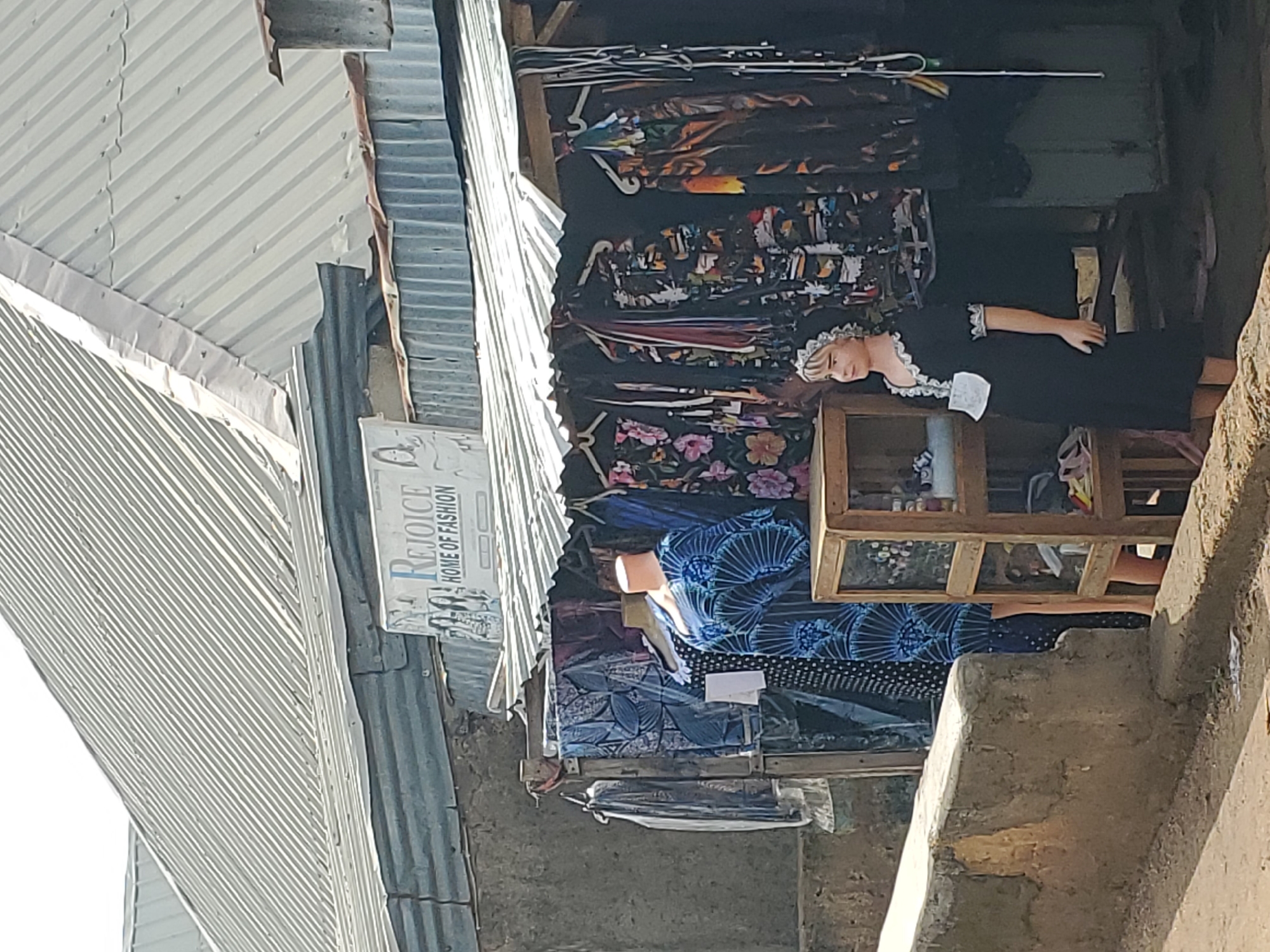 The image depicts an outdoor stall in Jos, Nigeria, specializing in fashion. A corrugated metal roof covers the stall, which displays various garments on hangers and mannequins. One mannequin, positioned to the right, is adorned in a black dress with white lace trim and a white headpiece. To the left, two headless mannequins are dressed in brightly patterned blue and white dresses. Behind the mannequins, a variety of dresses and fabrics with intricate patterns are visible, including floral designs. A wooden display shelf is situated in front of the mannequins, holding some smaller items. The stall is built against a rough, grey concrete wall. The overall setting suggests a bustling market environment in Jos.