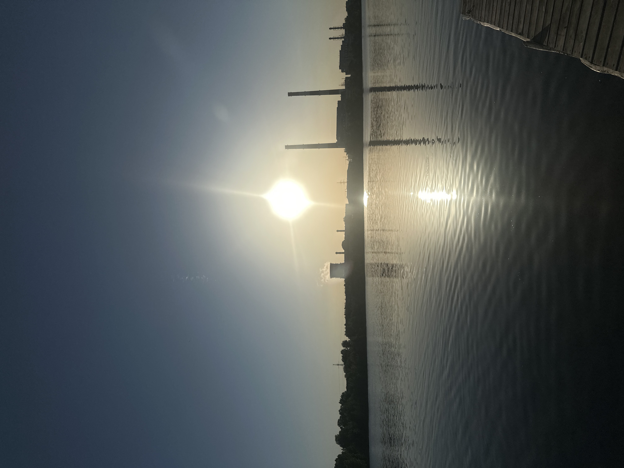 The image shows a body of water, likely a river or lake, with industrial structures silhouetted against the setting sun. Reflections of the sun and the structures are visible on the water's surface. The right edge of the frame shows part of a wooden dock or pier. The location is Kazan, Russia, as evidenced by the industrial structures which match a power plant on the outskirts of the city. The sky transitions from dark blue on the left to a brighter, sunlit haze near the horizon. There are a few wispy clouds.