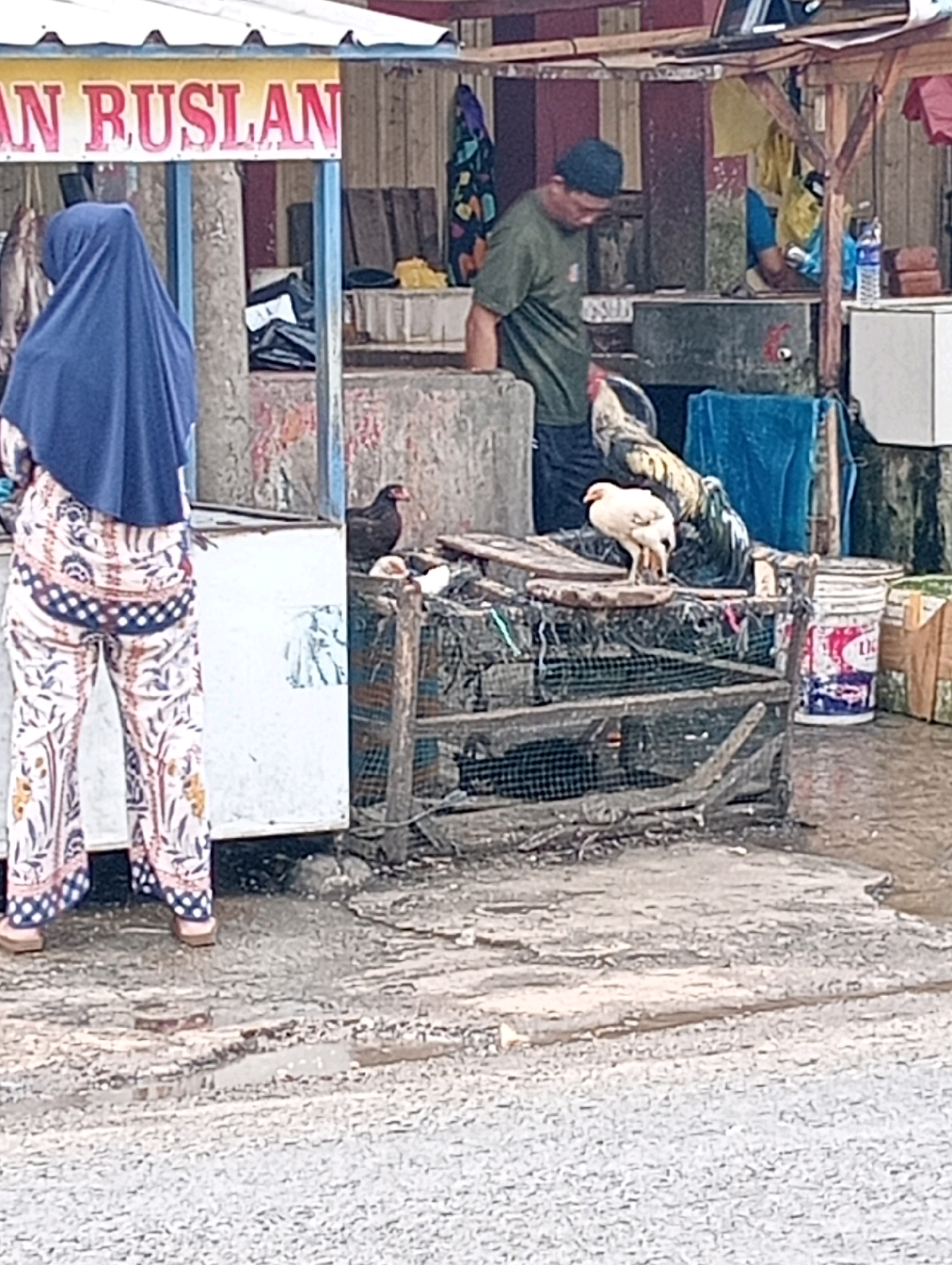 The image depicts an outdoor scene, possibly a market stall, with a woman in a blue hijab and a patterned outfit standing to the left, facing away from the camera. She appears to be near a stall with a sign that reads "AN RUSLAN". To her right, a man wearing a green shirt and a dark cap is visible, looking down. In the foreground, a wire cage contains several chickens. One black chicken is perched on the edge of the cage, and a lighter-colored chicken stands on a wooden plank above it. In the background, there are various items, including buckets, what appear to be fabrics, and a white structure. The ground is wet and uneven, with puddles indicating recent rain or damp conditions.