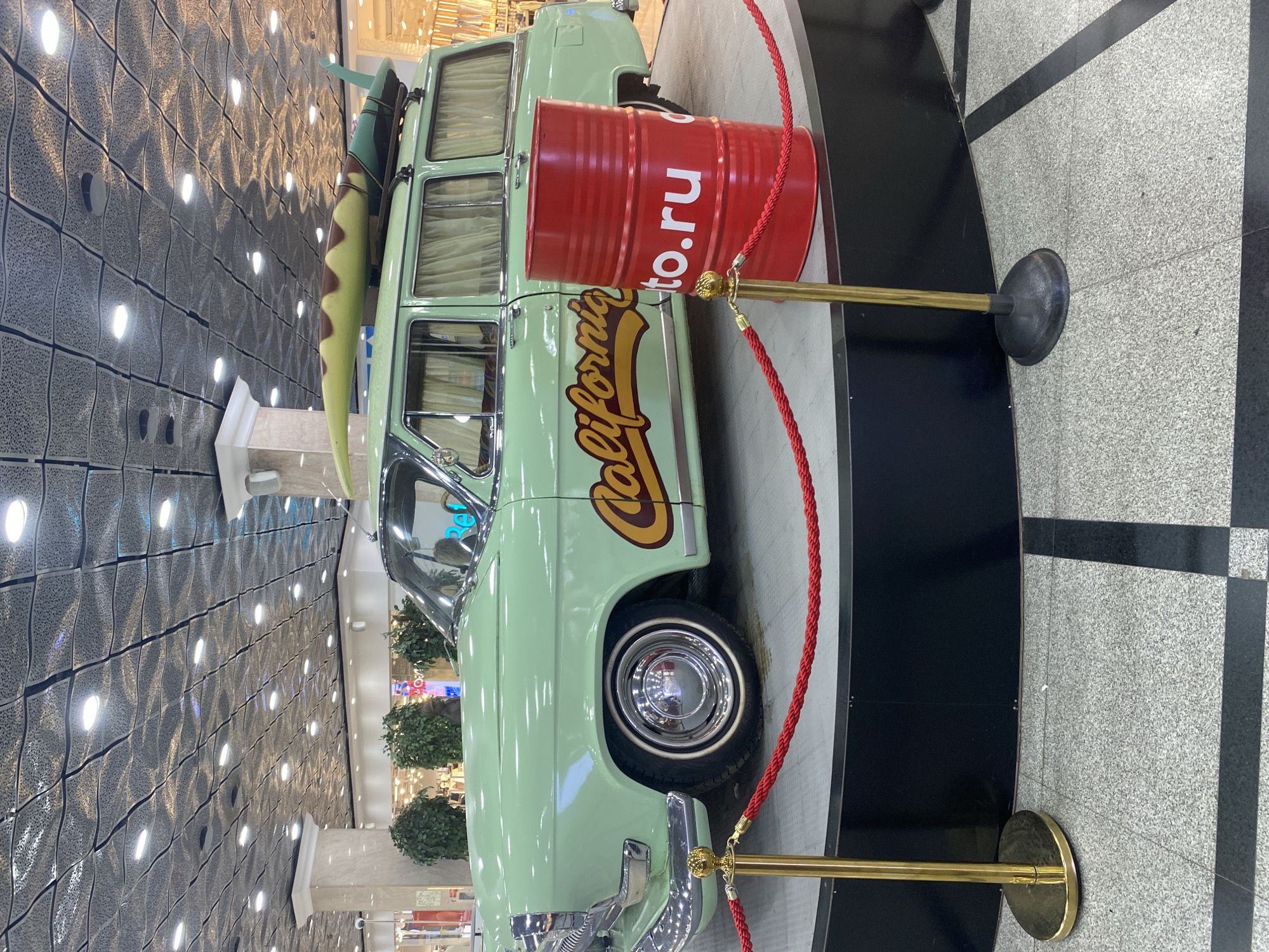 A light green station wagon with "California" lettering on the side, a surfboard strapped to the roof, and a red barrel with the text "auto.ru" displayed on the side is displayed on a black platform. The car is roped off with red rope and gold stanchions. The background shows a commercial interior with a decorative wall and several stores. The scene is located in the city of Yekaterinburg, Russia.