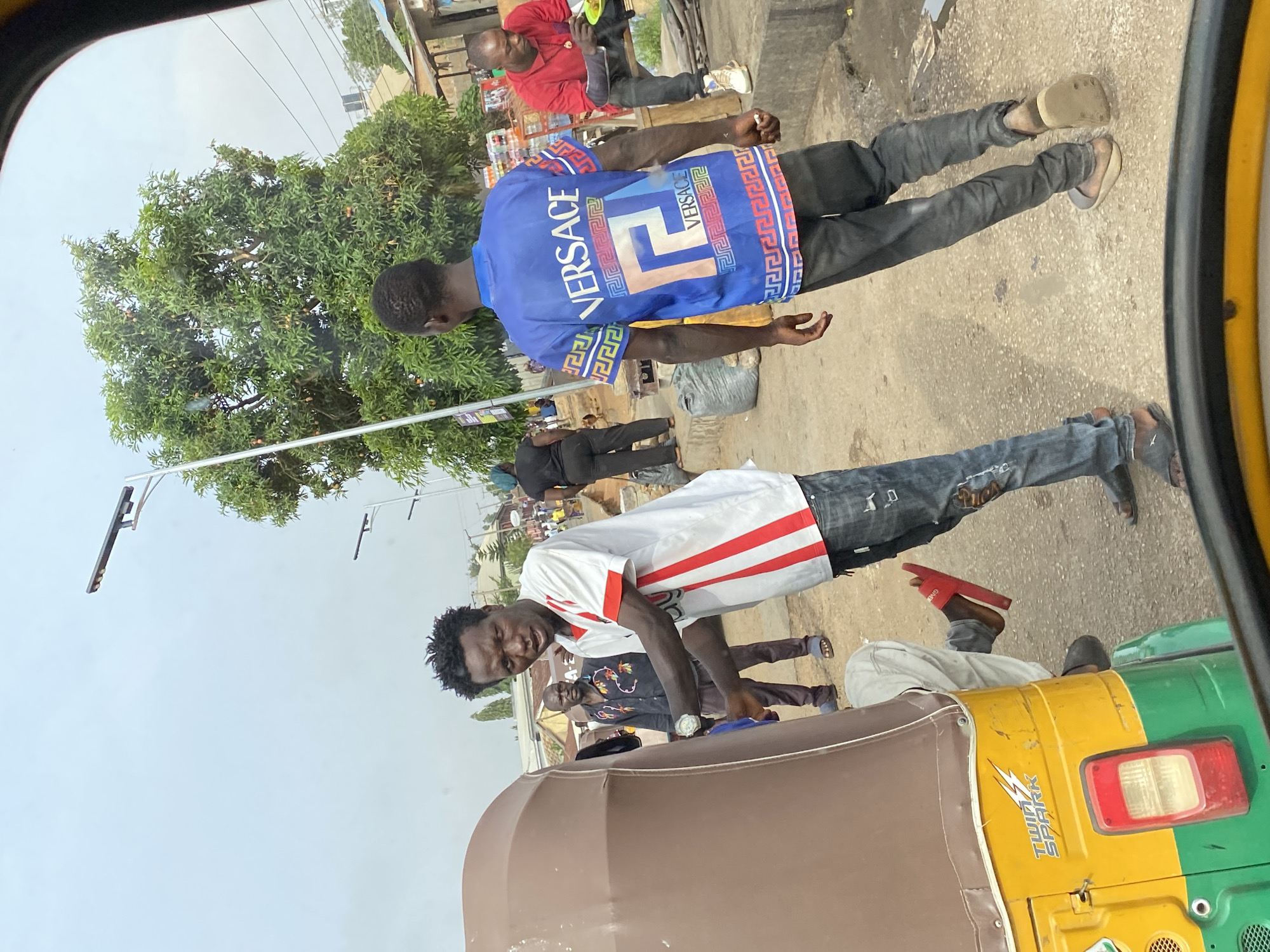 The image shows a street scene in Bukuru, Nigeria, viewed from inside a vehicle. Two men in distinct shirts are prominently displayed: one in a white shirt with red stripes and the other in a blue "VERSACE" shirt. Other individuals are scattered in the background. A yellow and green motorized tricycle is visible on the right side of the frame. There are streetlights and trees also present in the frame.