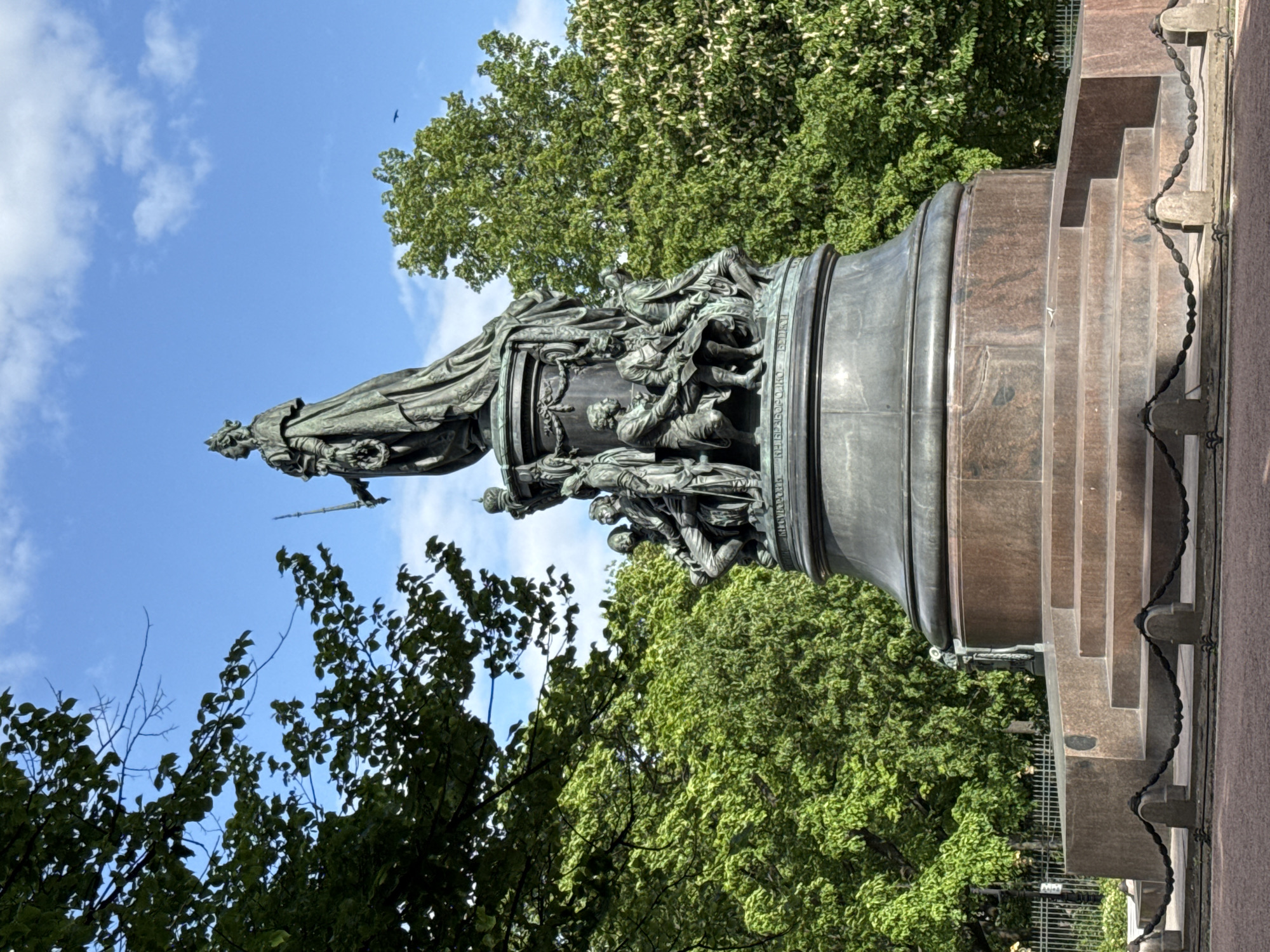The image captures a view of the Monument to Catherine the Great in Saint Petersburg, Russia. The bronze monument stands prominently against a backdrop of green trees and a partly cloudy sky. The statue of Catherine is visible at the top, and various figures are sculpted around the base of the column. The monument is set upon a red marble base with several steps, and a section of a fence with hanging chains is visible to the right of the monument.