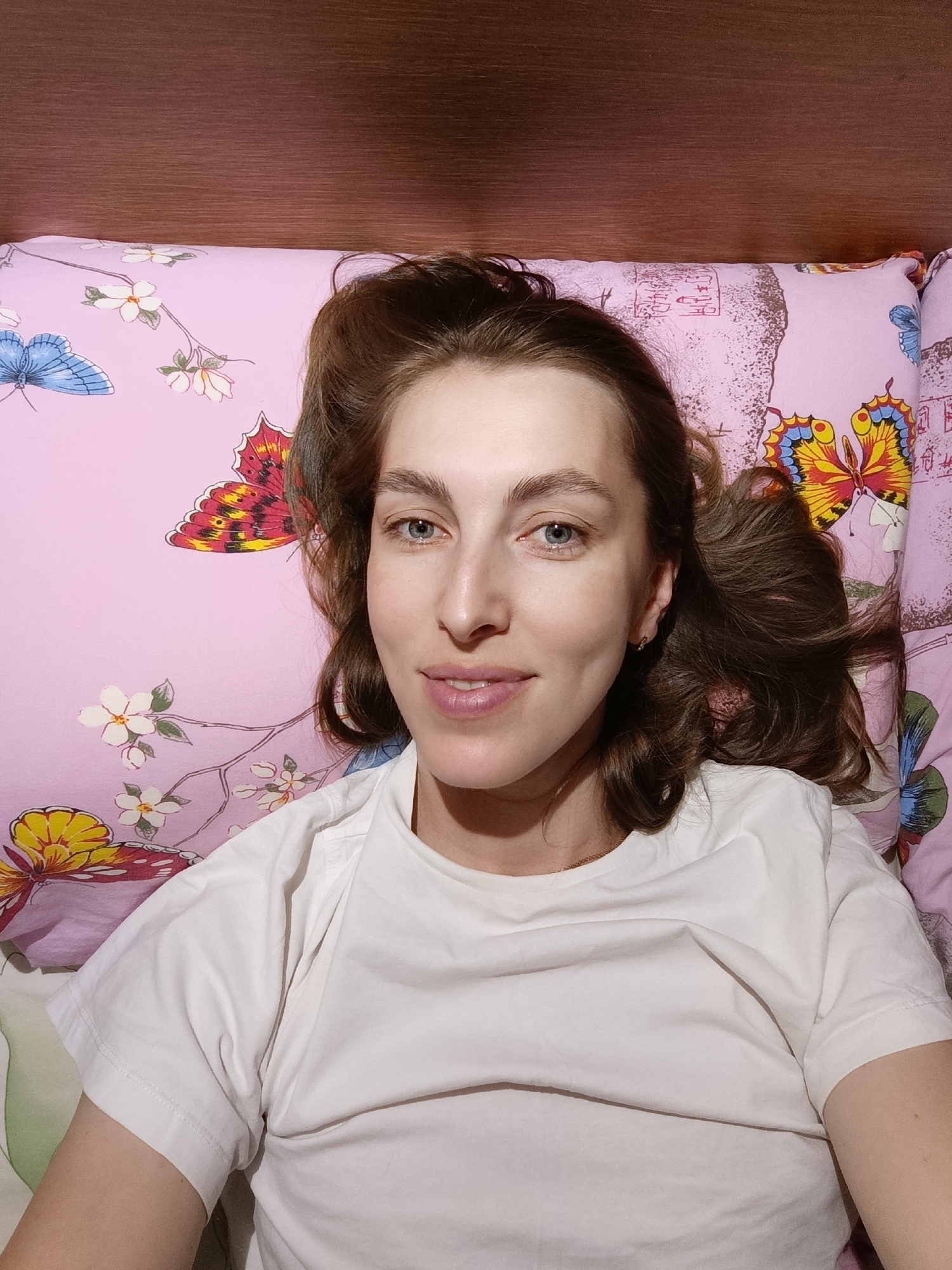 A young woman with brown hair and blue eyes lies on a bed, looking up at the camera. She is wearing a white t-shirt and her head is resting on a pink pillow with a butterfly and floral pattern. A wooden headboard is visible behind the pillow. The image appears to be a selfie, taken from a slightly elevated angle. The background suggests an indoor domestic setting. The image was likely taken in Chaikovsky, Russia.