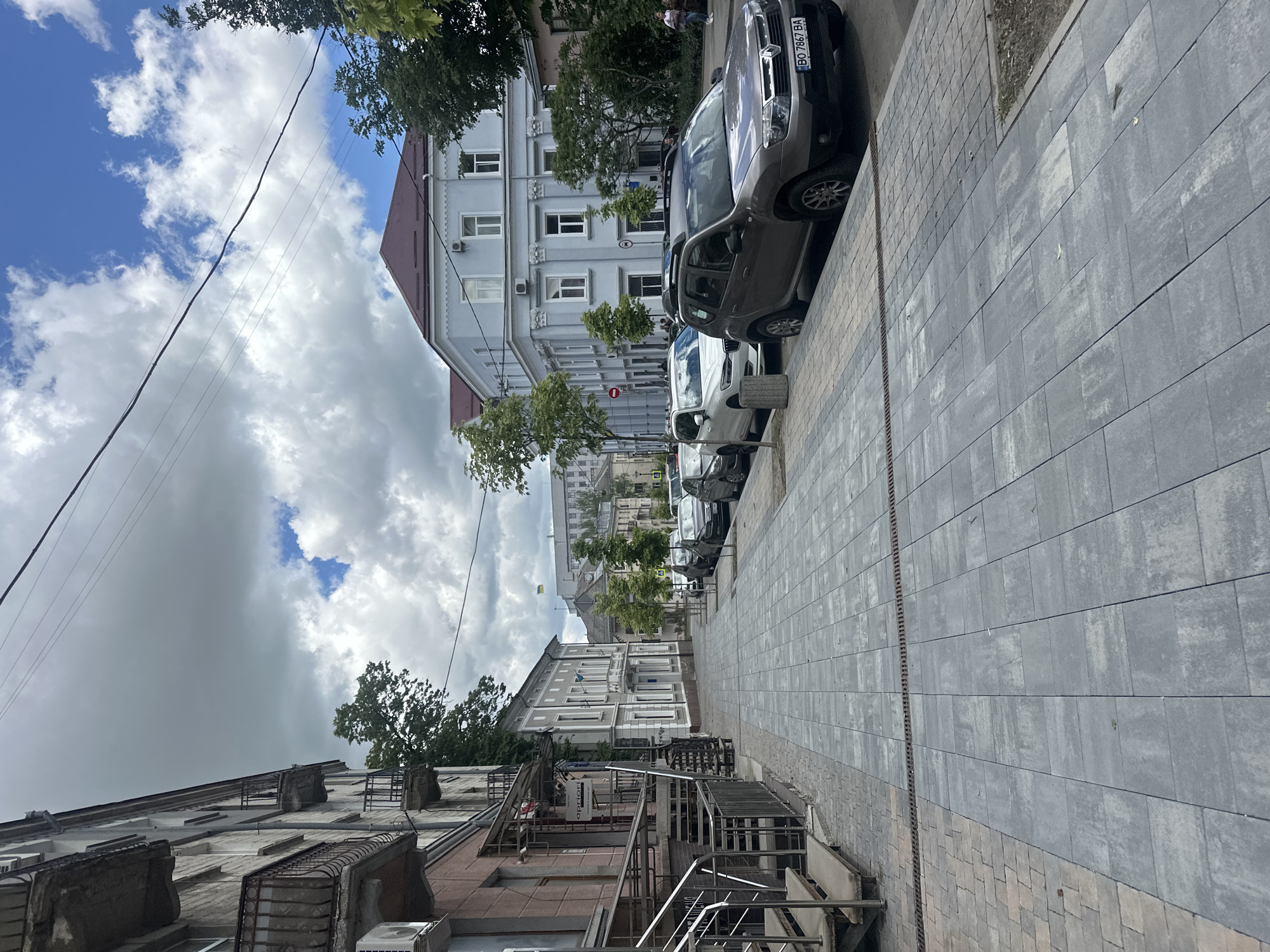 This is an outdoor shot in the city of Ternopil, Ukraine. The image is oriented vertically, depicting a street scene with buildings lining both sides.
On the right side of the image, several cars are parked along the edge of a gray-tiled sidewalk. The first car in the row is a dark-colored SUV, with the partial license plate "BO 7867 BA" visible. The sidewalk itself is composed of large gray rectangular pavers.
On the left side, a building with a pinkish-brown facade is visible. It has metal staircases and balconies. Across the street, a light gray building with detailed window trims can be seen. Trees are interspersed along the street, partially obscuring the views of the buildings.
The sky above is filled with a mix of blue patches and thick, white and gray clouds. Overhead power lines are visible against the sky. The perspective of the image indicates a street that stretches into the distance, with buildings becoming smaller.