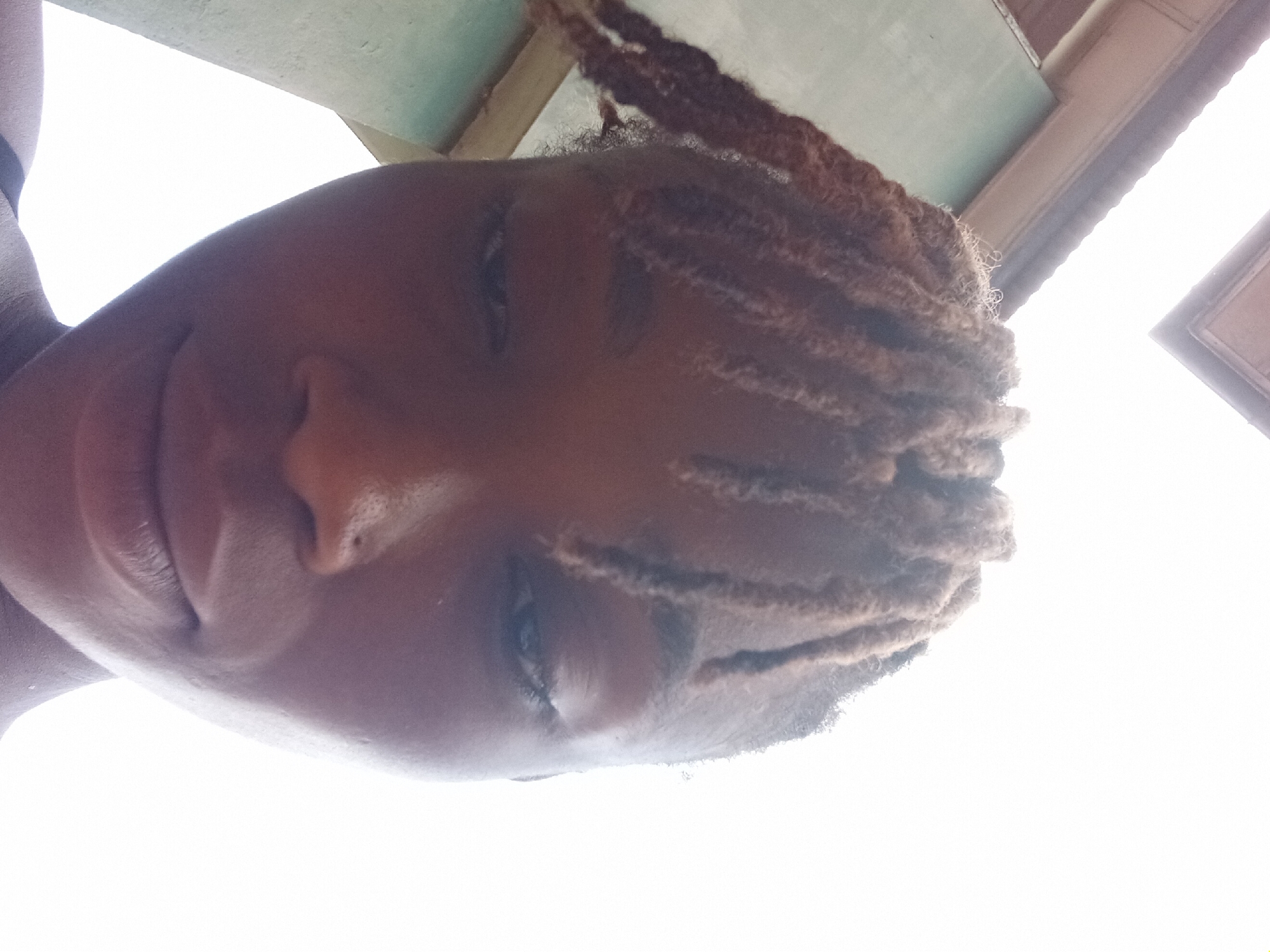 A person with dreadlocks is seen from a low angle, with a light-colored building or structure in the background. It is daytime, and the location is Shagamu, Nigeria.