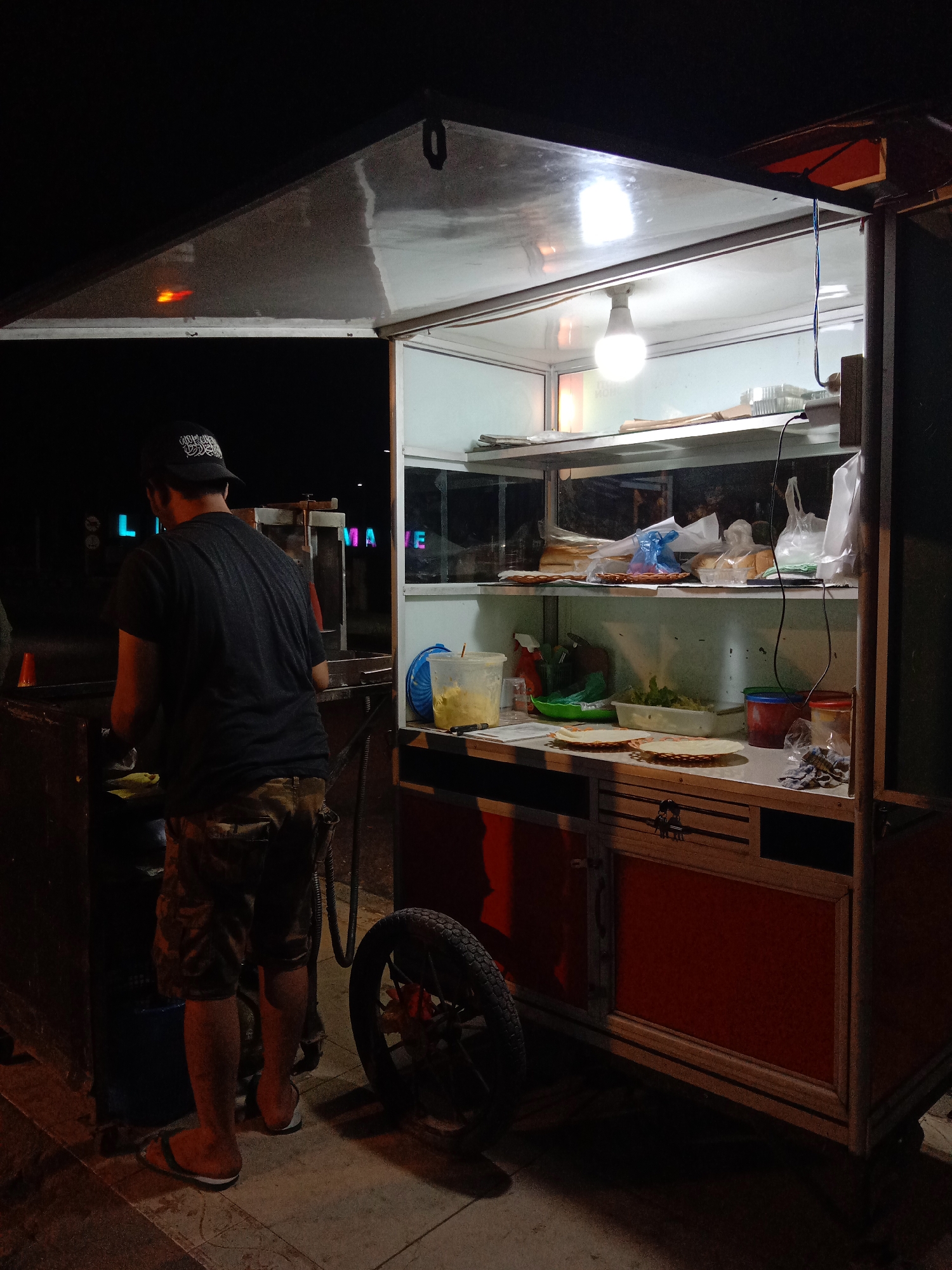 The image captures a food cart vendor operating at night in the city of Lhokseumawe, Indonesia. A man, wearing a black t-shirt and camouflage shorts, stands behind the cart, seemingly preparing food. The cart itself is illuminated by a single bright bulb, casting light on shelves stocked with various food items and packaging, including what appear to be bread, vegetables, and sauces. A blue cooler is visible on the left side of the cart, and a wheel is attached to the right. In the background, neon signs spell out "L HOKSEUMAWE," indicating the location. The scene suggests a typical street food setup, with the vendor actively engaged in their business under artificial light against the dark night sky.