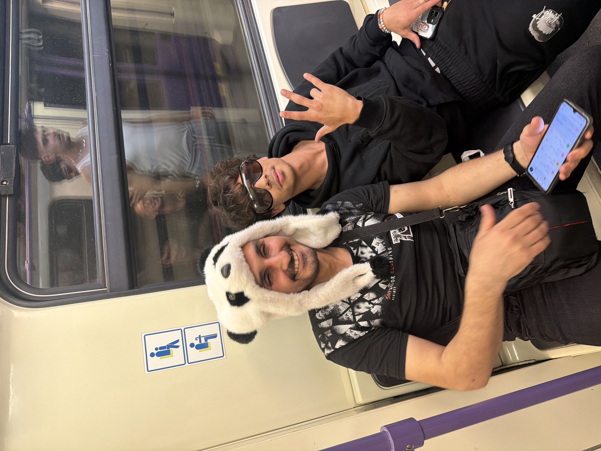 Two young men are reclining on seats in a train car. The man in the foreground is wearing a panda hat and a black t-shirt with a design, holding a black smartphone with a colorful display. He's wearing a black watch and a black shoulder bag. The man behind him is wearing sunglasses and a black hooded sweatshirt. He is holding a smartphone in his hand and making a hand gesture with his other hand. Their reflections are visible in the window. There is a sign on the wall with stick figures indicating seats for people with disabilities and pregnant women. The color scheme of the train interior is light beige with purple accents. It's a carriage in the metro of Sofia, Bulgaria.