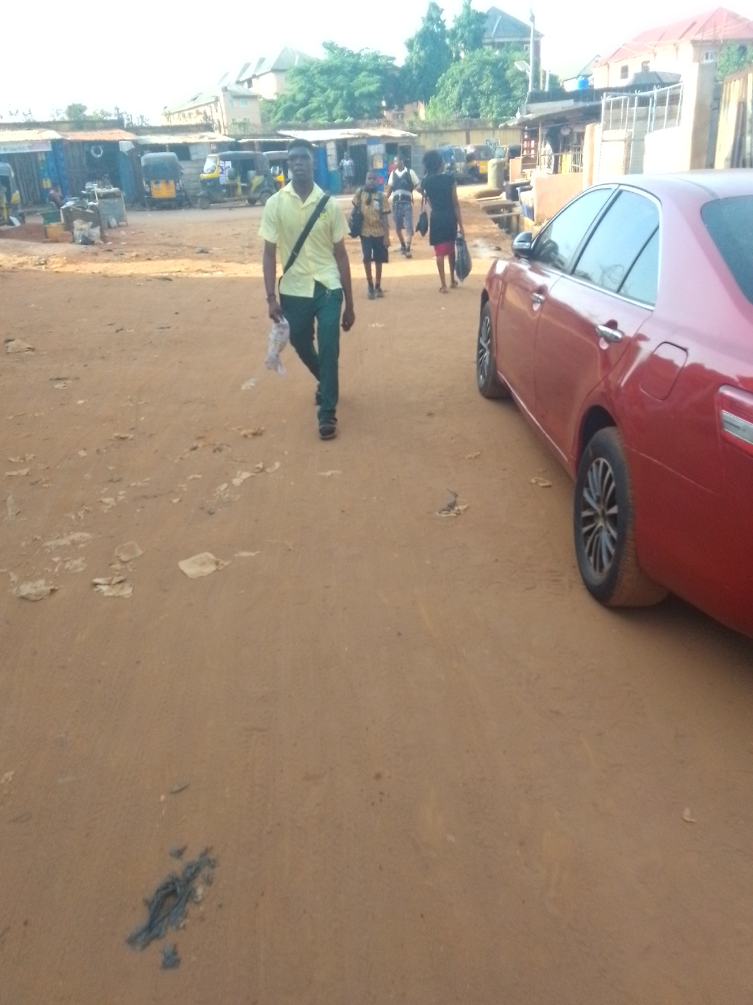 The image depicts an unpaved street in Onitsha, Nigeria. A young man in a yellow shirt and dark trousers walks towards the camera, carrying a plastic bag. Behind him, two other individuals are walking in the same direction. A red sedan is parked on the right side of the frame. The street surface is rough, with scattered debris. In the background, there are shops and buildings. Tricycles are visible, parked along the side of the street.