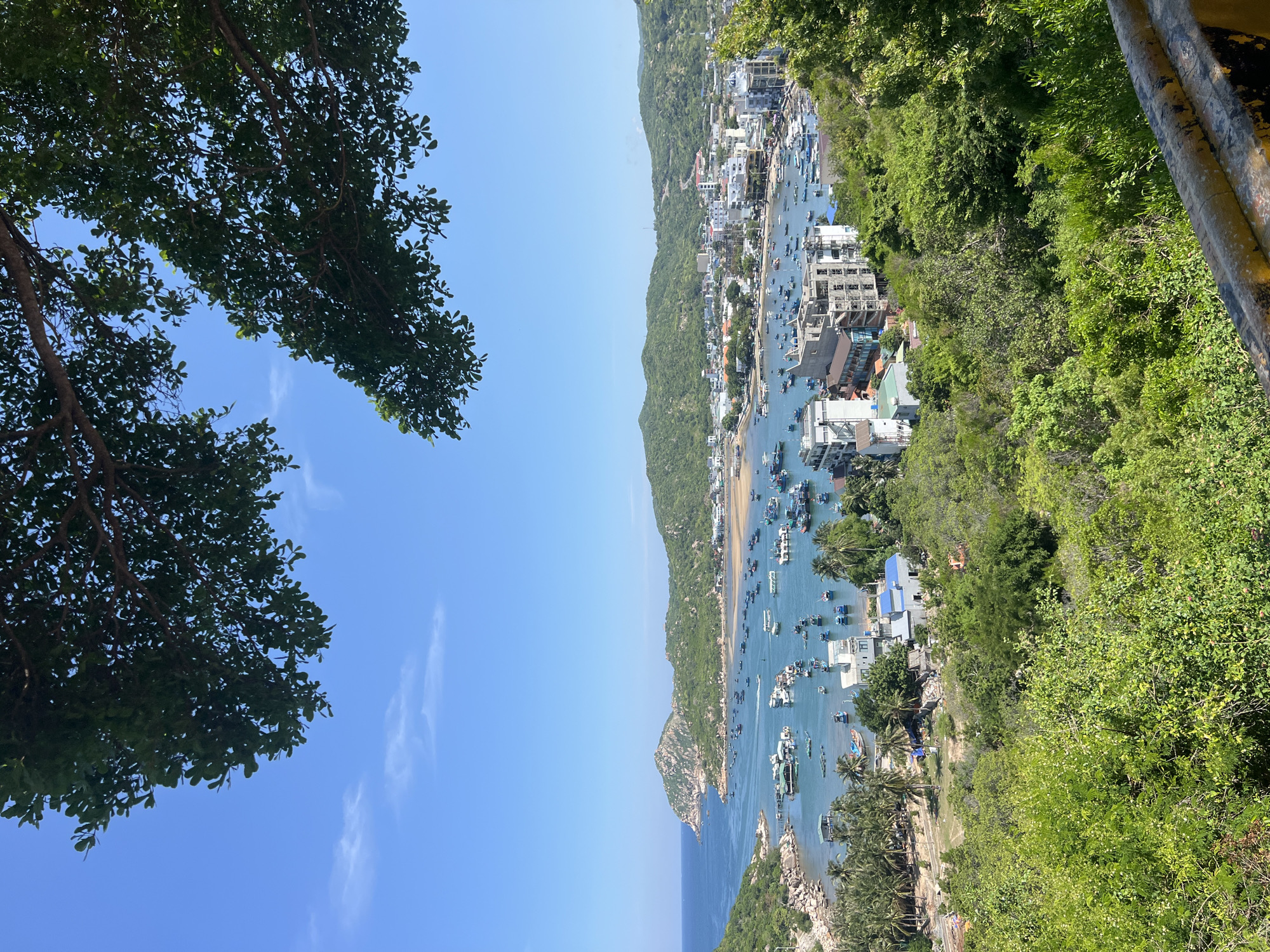 A high-angle, full shot captures a vibrant coastal town nestled in a bay. The sky is a clear, bright blue, with scattered white clouds. In the foreground, lush green trees frame the scene from the top, and a yellow-and-black metal barrier runs along the bottom right.
The bay itself is dotted with a multitude of boats, varying in size and color, anchored in the turquoise water. These boats range from small, traditional fishing vessels to larger, more modern craft. Some appear to be docked near a pier or floating structures.
Along the shoreline of the bay, a town sprawls, characterized by a mix of buildings. There are low-rise structures, some appearing to be residential homes, and several taller buildings, including multi-story hotels or apartment complexes. Some buildings are under construction, with exposed concrete frameworks.
The town is backed by rolling green hills covered in dense vegetation, creating a picturesque backdrop. The coastline curves around the bay, with a rocky outcrop visible on the left side, also covered in greenery. The overall impression is a bustling coastal community during daylight hours, likely a popular tourist destination within Vĩnh Hải, Vietnam.