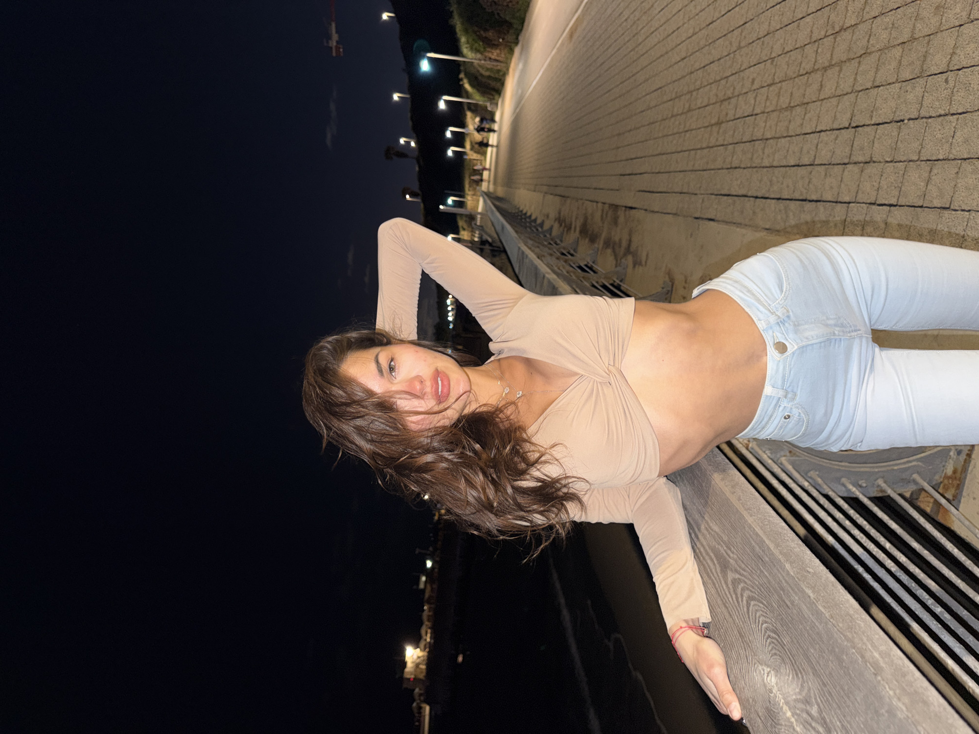 A female subject stands on a paved promenade at night in the city of Unknown, Unknown. She wears a light beige long-sleeved crop top with a twisted front and light blue jeans. Her dark, wavy hair is partially windblown, and her left hand is placed behind her head while her right hand rests on a wooden and metal railing. The subject looks directly at the camera. To her left, beyond the railing, is a dark body of water with illuminated structures visible on a distant shoreline. To her right, a paved walkway, constructed from diamond-patterned pavers, extends into the background, illuminated by multiple tall streetlights. Several indistinct human figures are visible walking in the distance on the pathway. A vegetated slope borders the walkway, also featuring streetlights. A red and white construction crane is partially visible in the upper right background against the dark, sparsely clouded night sky.