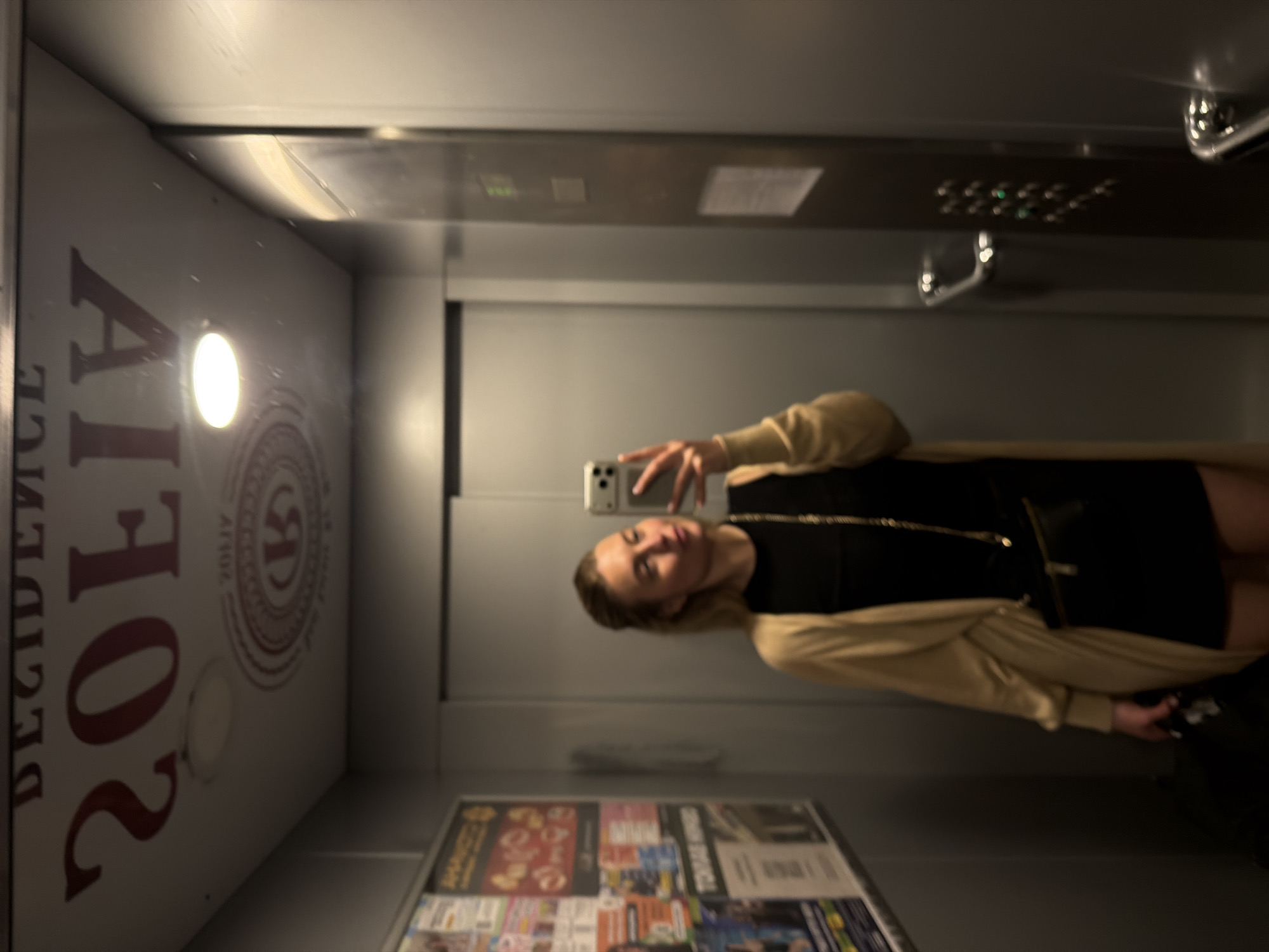 A female individual is captured in a mirror selfie inside an elevator located in Sofiivska Borshchahivka, Ukraine. The subject, positioned centrally, is a woman with light hair, wearing a black garment, a long beige cardigan, and a gold-colored chain. She holds a silver smartphone with both hands, pointing its rear cameras towards the mirror for the reflection. A dark fanny pack or waist bag is worn around her hips.
The elevator's interior features grey walls with reflective surfaces. The ceiling displays mirrored dark red text and a circular emblem. When un-mirrored, the text reads "RESIDENCE" on an upper line and "VILIOS" in a larger font below, with "VILIOS" also present in smaller script within the circular logo. A single circular recessed light fixture on the ceiling provides illumination.
On the left wall (from the viewer's perspective), a metal-framed notice board contains multiple overlapping posters and flyers, primarily with Cyrillic text that is not legible. On the right, a grey elevator control panel is visible, featuring a vertical array of buttons, some illuminated green, and a faint green digital display showing a "2" or "3" (likely indicating the current floor). Two polished metal grab handles are affixed to the control panel. The floor of the elevator is dark. The elevator doors, reflected behind the subject, are flat and grey.