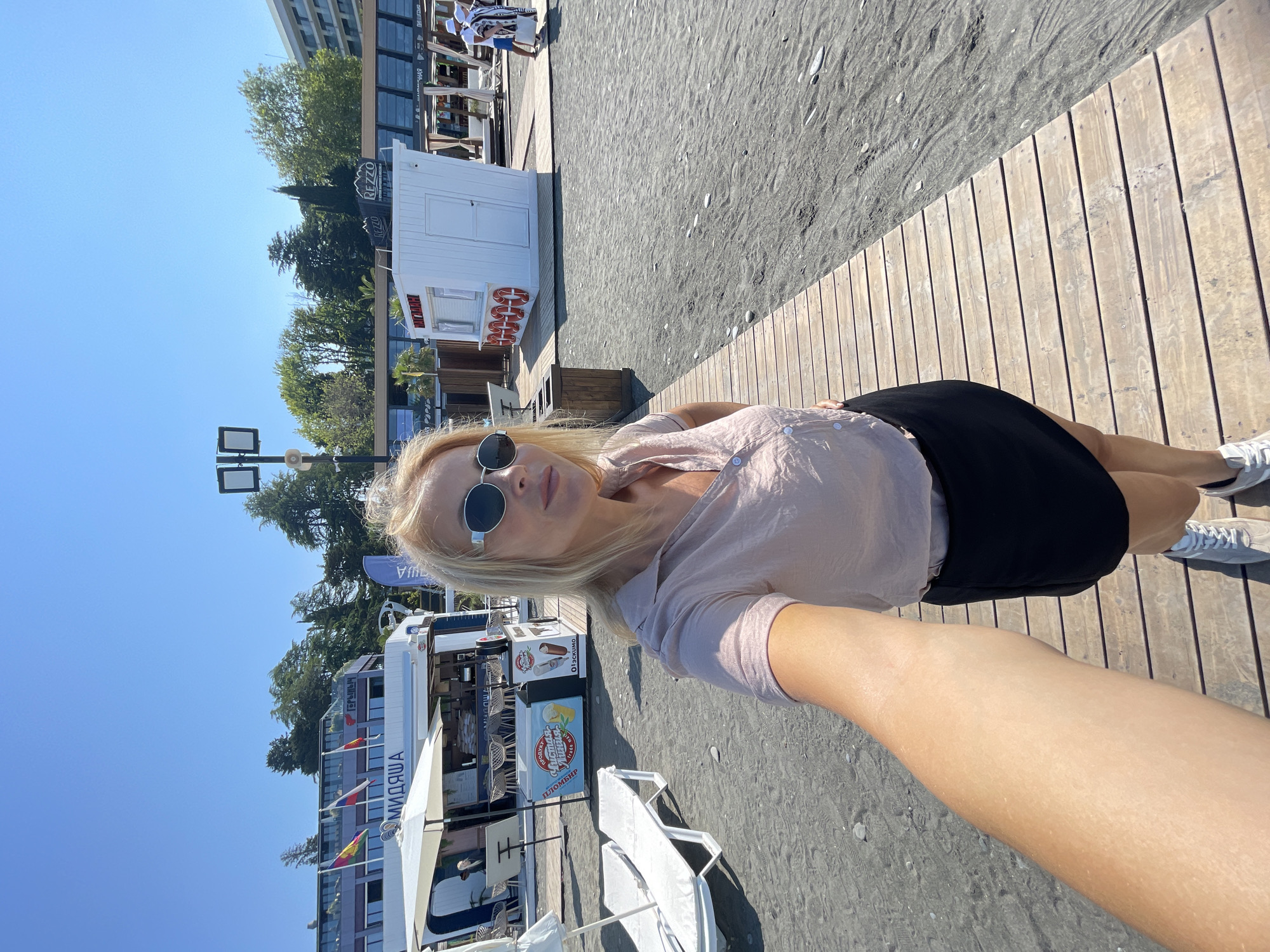 A blonde woman is taking a selfie on a wooden boardwalk on a beach. She is wearing sunglasses, a light pink shirt, and a black skirt, and white sneakers. The boardwalk leads towards the sea, which is not visible in the frame. In the background, there are beach chairs, a food kiosk, and buildings. The sky is clear and blue. The beach is covered in dark sand and small pebbles.