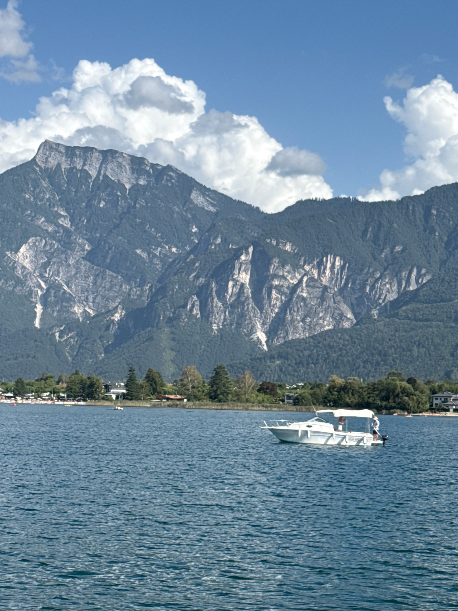 This is a vertical, eye-level shot of a wide lake with a boat and mountains in the background. The lake's surface is choppy blue, with whitecaps reflecting the sunlight. A white motorboat with a white canopy is in the right-middle of the frame. Two people, one wearing a white hat, are visible on the boat. In the far background, a range of imposing mountains dominates the scene. The mountains are a mixture of dark green forests at the base and sheer, rocky grey and white cliffs higher up. The sky above is a bright blue with scattered, puffy white clouds. Along the shoreline to the left of the boat, a strip of land is visible with trees and some buildings, suggesting a populated area or a beach. The scene appears to be the lake in Calceranica al Lago, Italy.